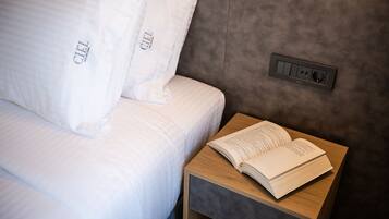 Una cama bien hecha con sábanas y almohadas blancas, una mesita de noche con un libro abierto y un enchufe en la pared.