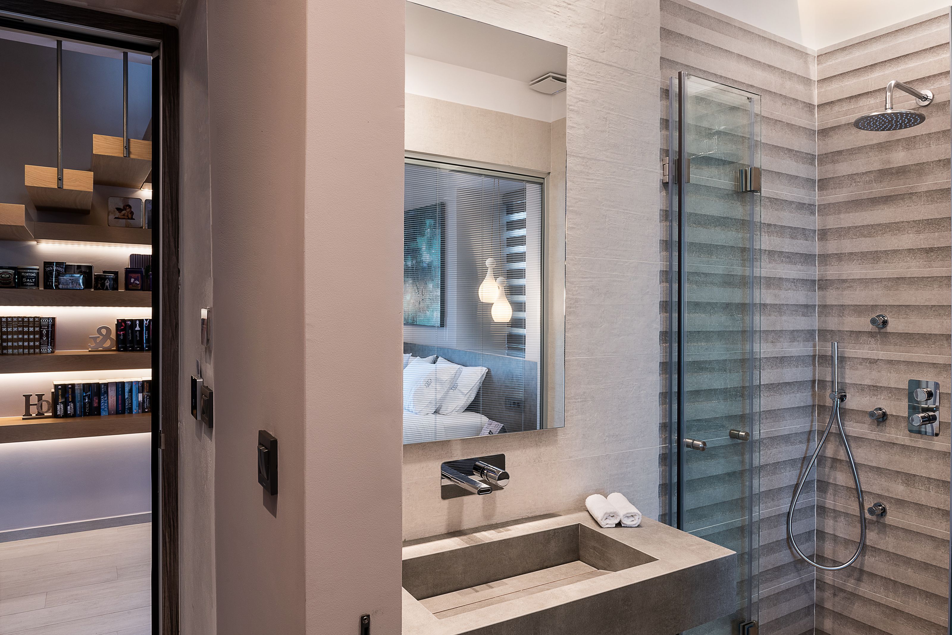 Een moderne badkamer met een douchecabine van glas, een inbouw wastafel en een grote spiegel die een slaapkamer weerkaatst.