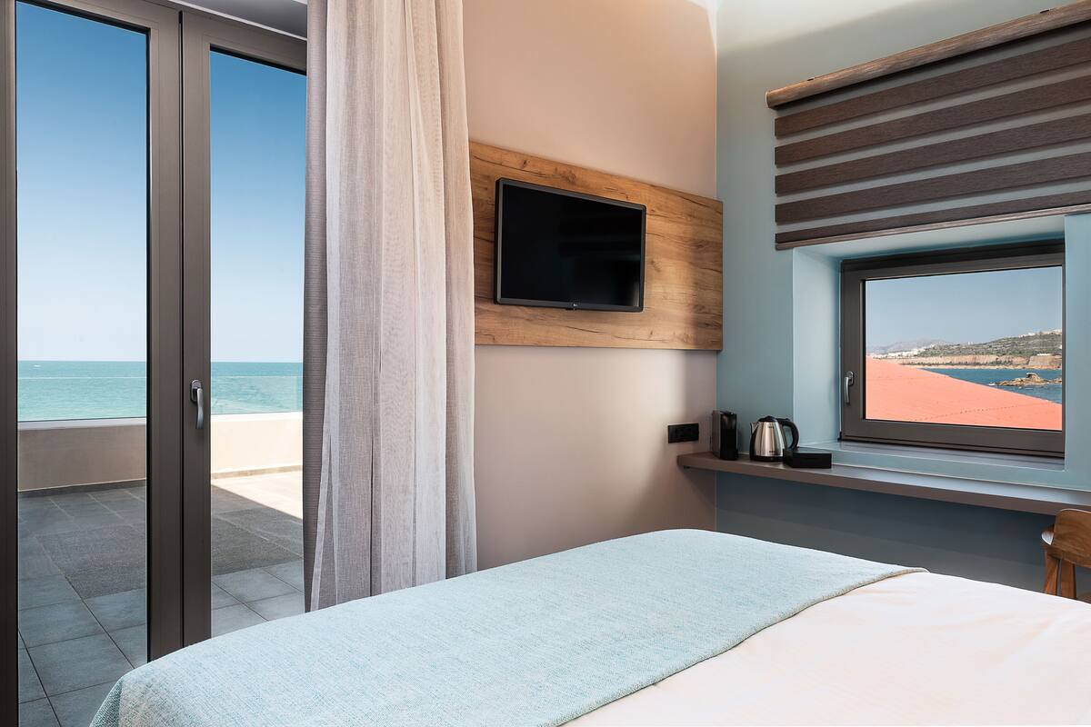 A bedroom with a bed, a TV mounted on the wall, a window with a view of the beach, and a small shelf with a kettle.