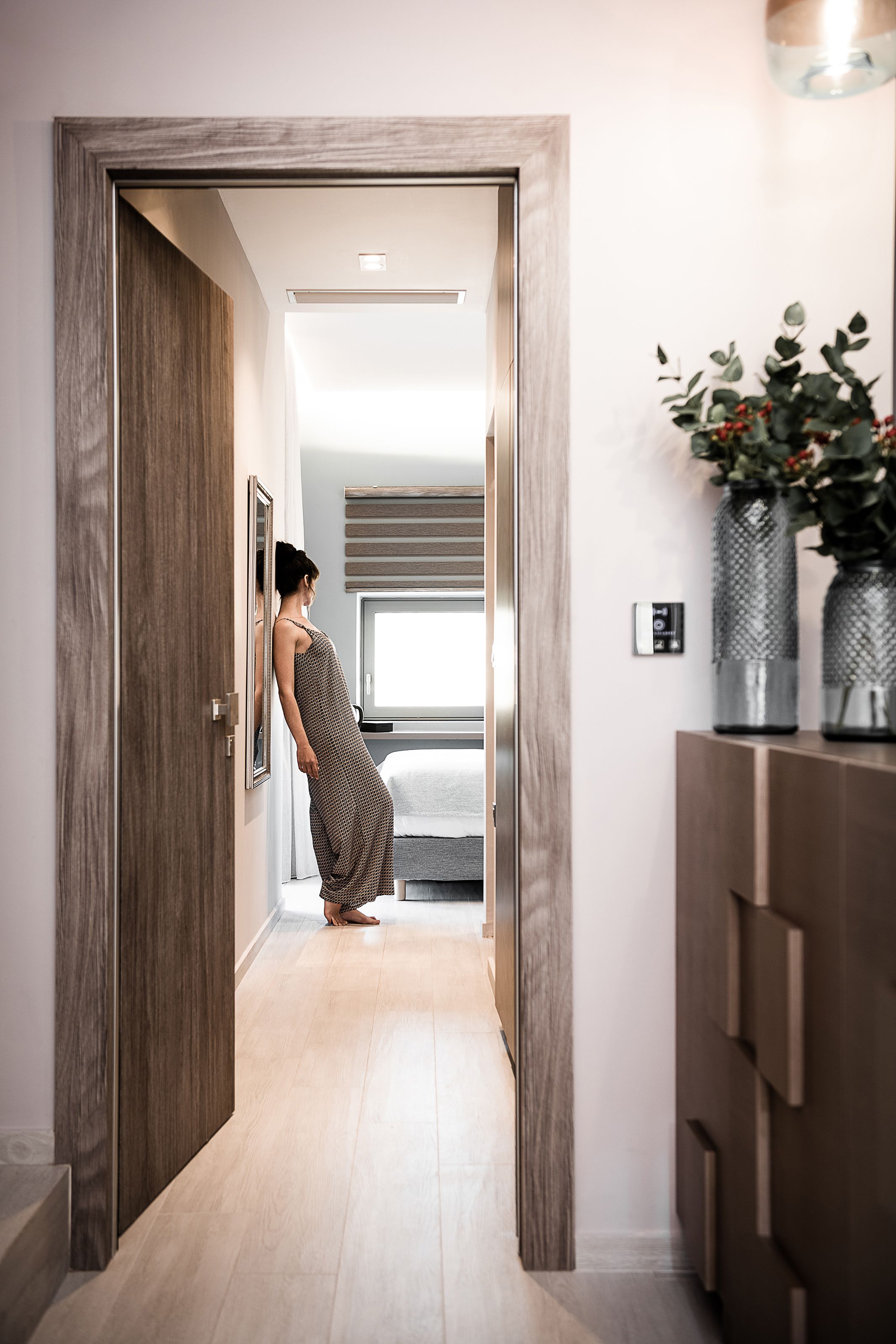 A hallway with wooden doors, a bed with a white headboard, and a vase with flowers on a cabinet.