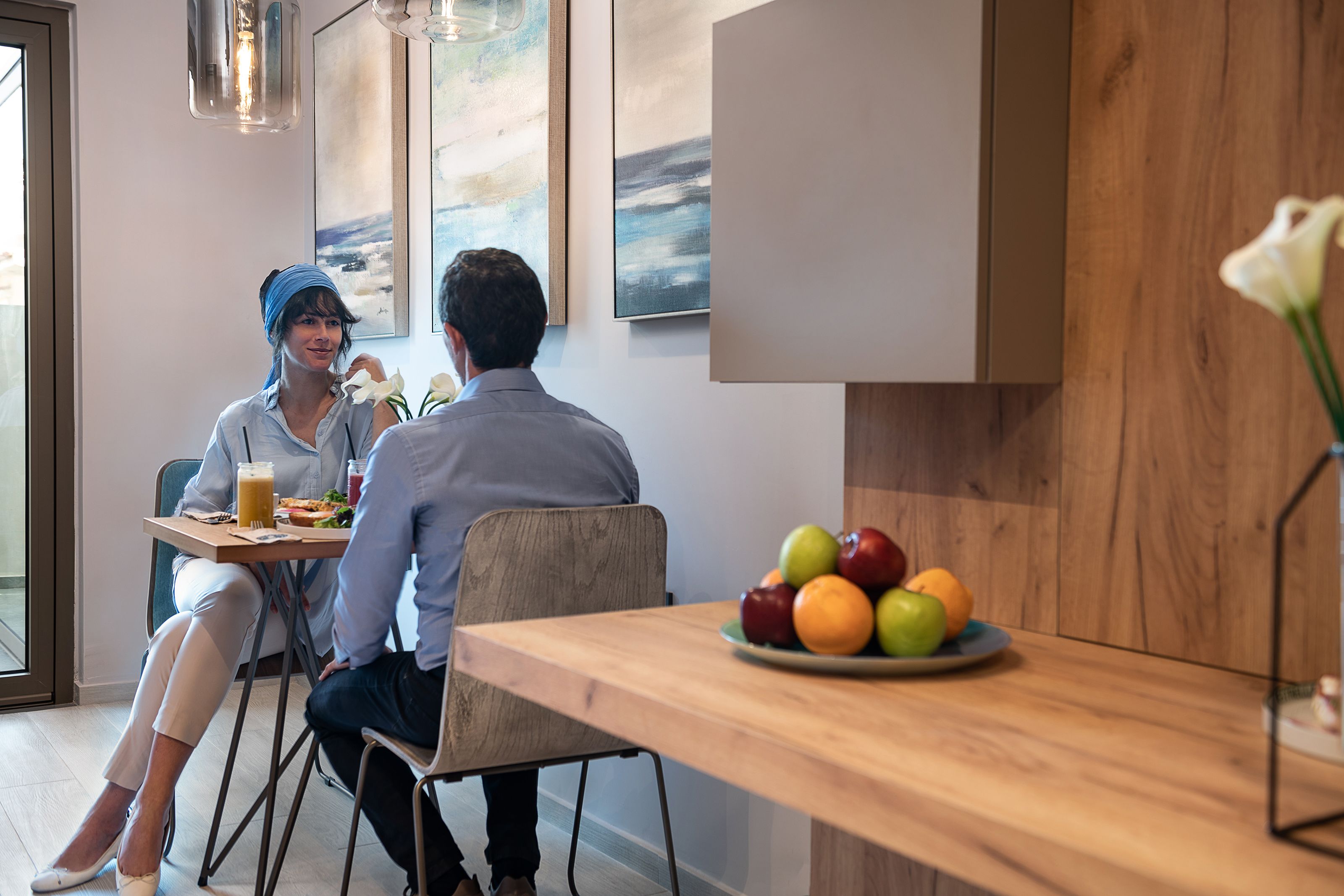 A modern dining area with a wooden table, a fruit bowl, and a painting on the wall.