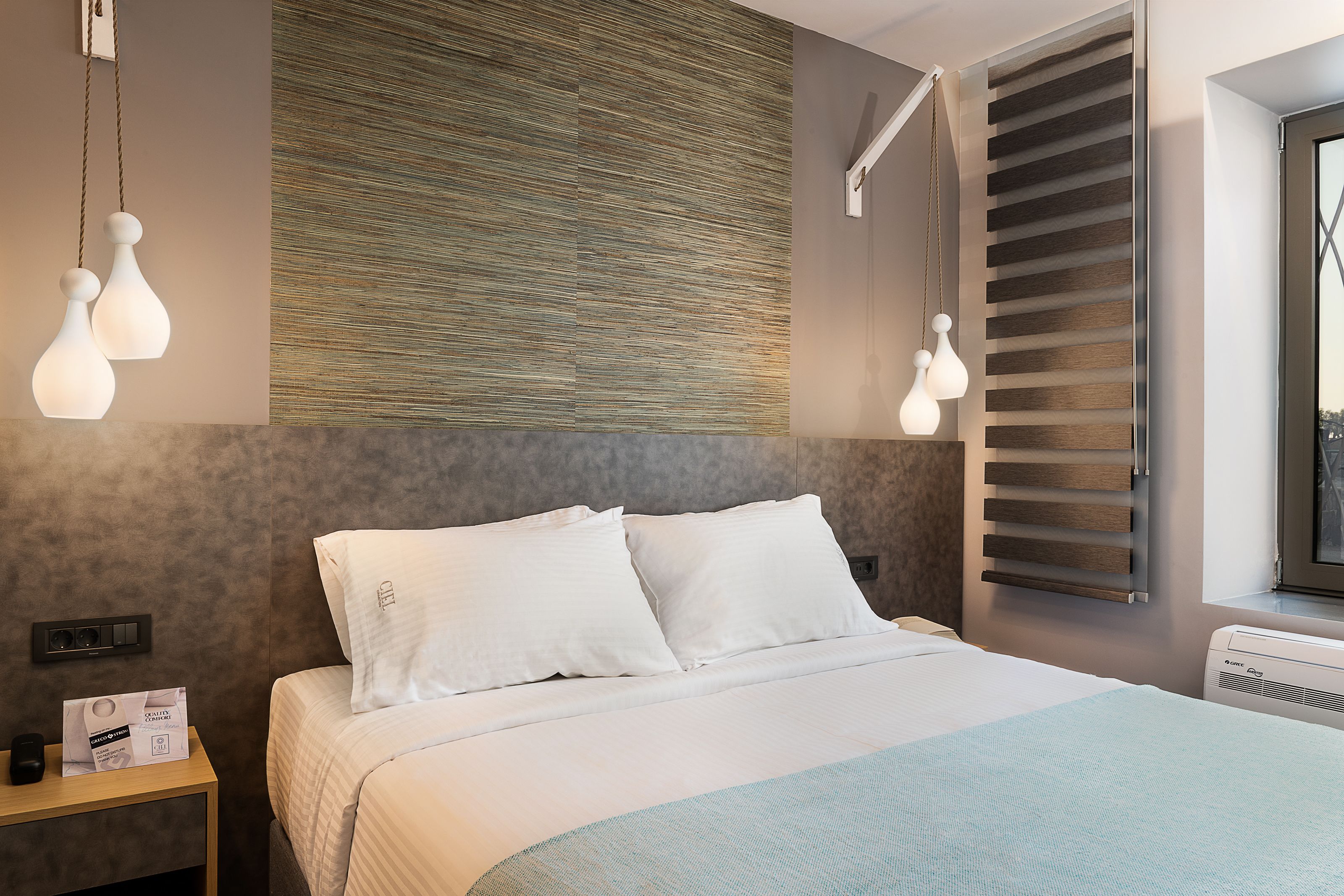 A modern hotel room with a large bed, a nightstand, a wall-mounted heater, and a window with blinds.