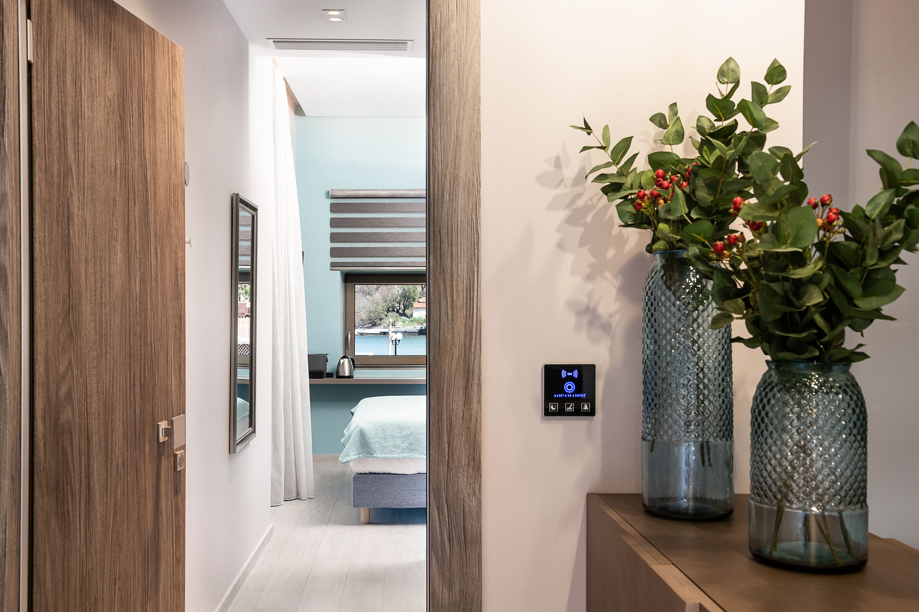 A hallway with a wooden door, a bed with a blue headboard, and two glass vases with green plants on a wooden shelf.