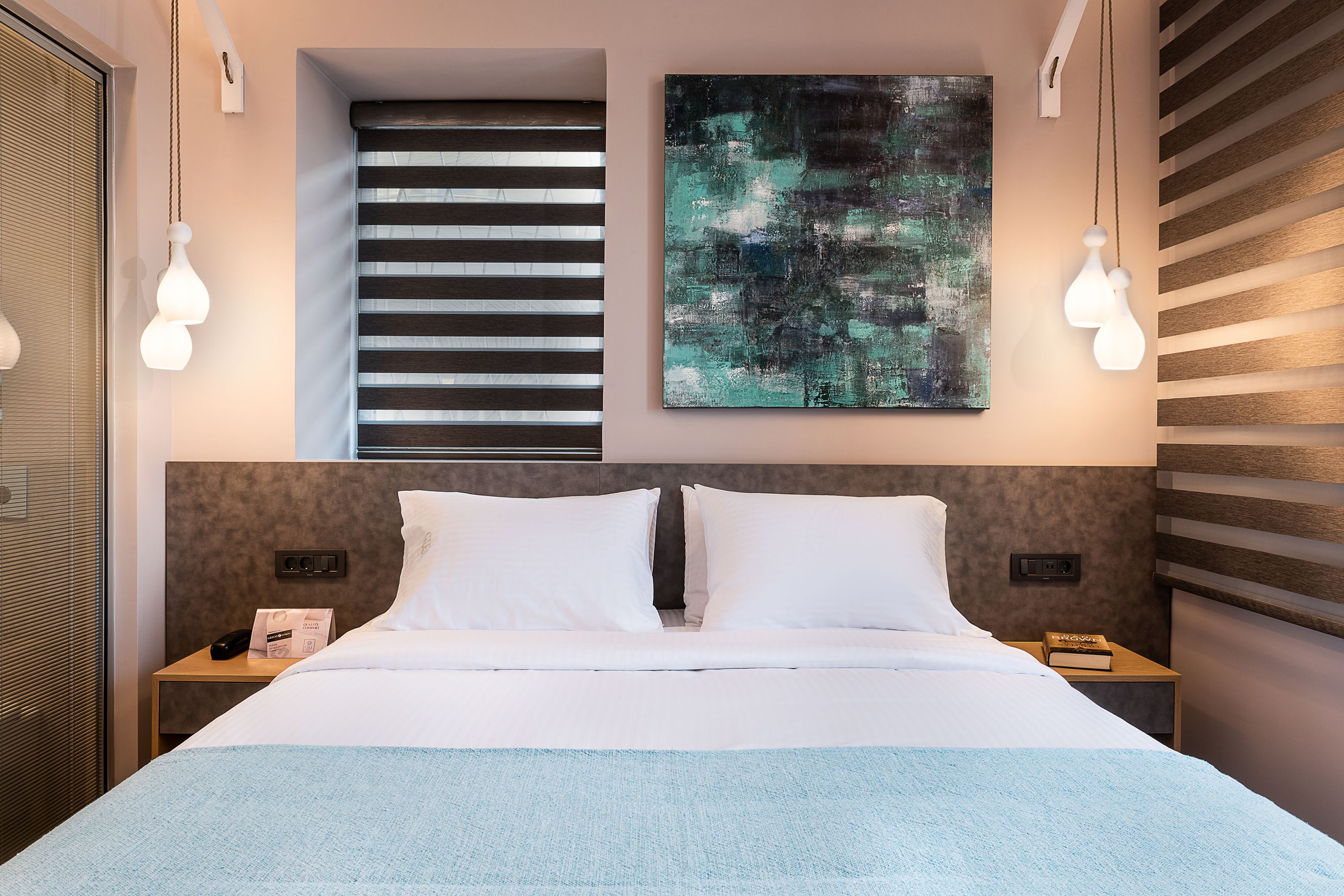 Une chambre d’hôtel moderne avec un grand lit, un tableau au mur et une fenêtre avec des stores.