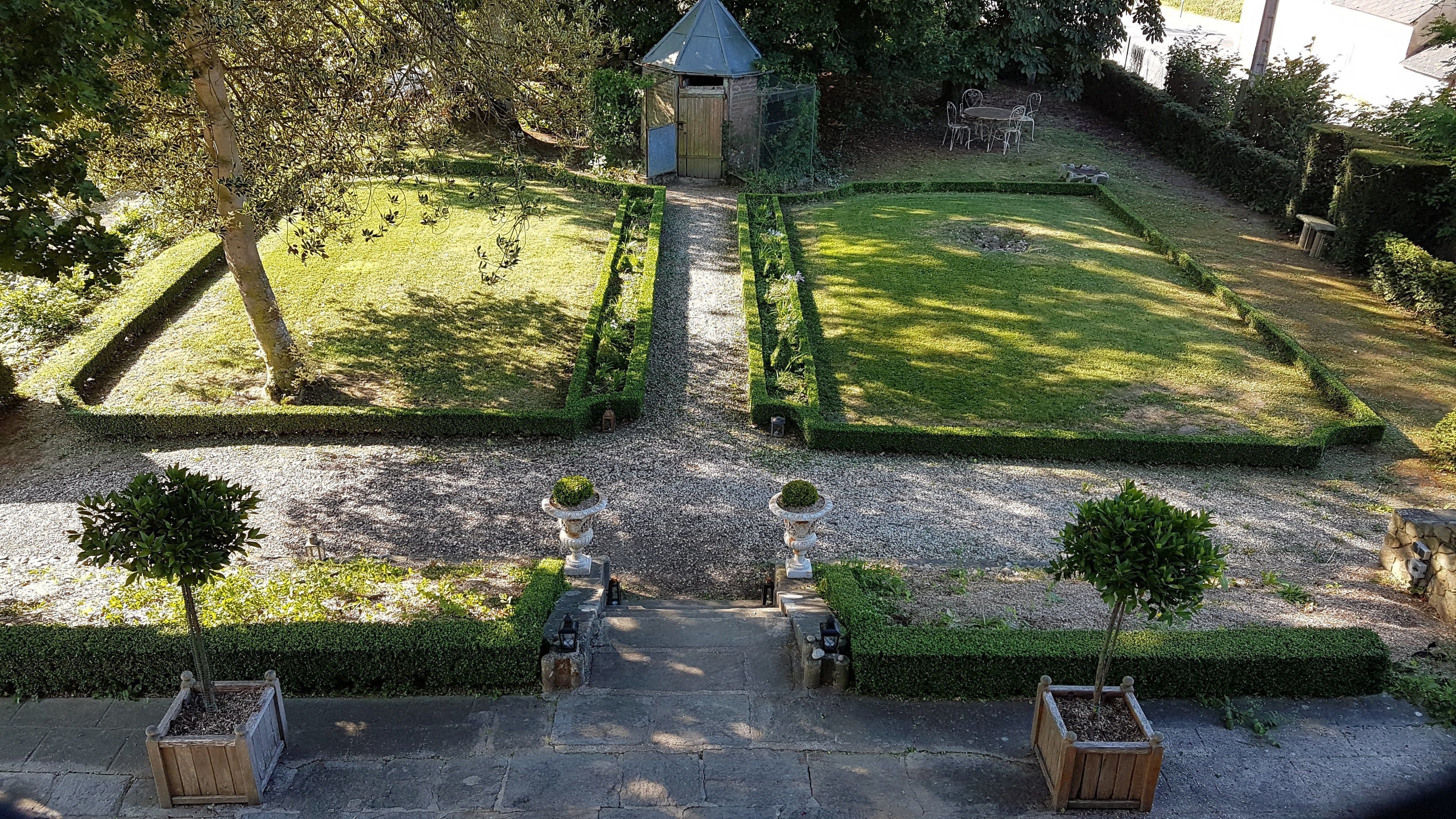 Ein Garten mit einem gepflasterten Weg, der zu einem Gazebo führt, umgeben von geschnittenen Hecken und kleinen Bäumen.