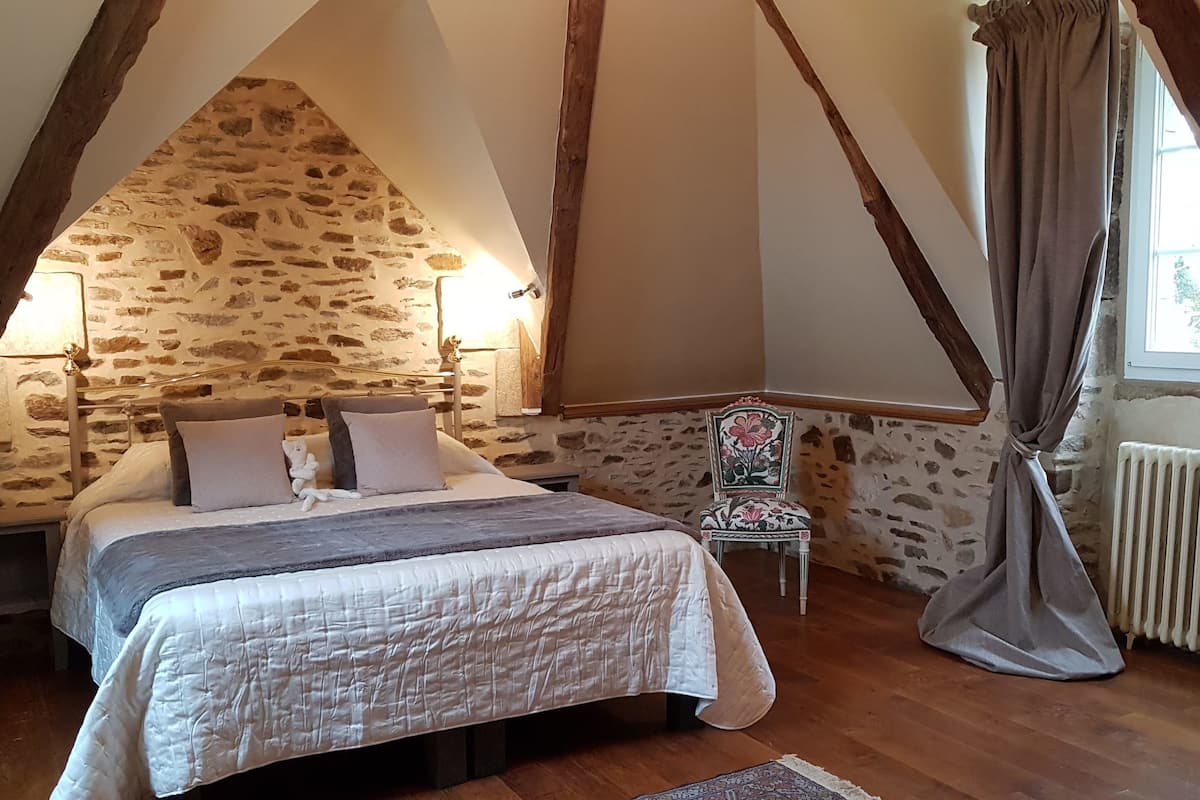 Un dormitorio con pared de piedra, una cama con almohadas, una silla y una ventana con cortinas.