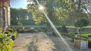 Un patio de piedra con una mesa y sillas, rodeado de setos y árboles bien cuidados.