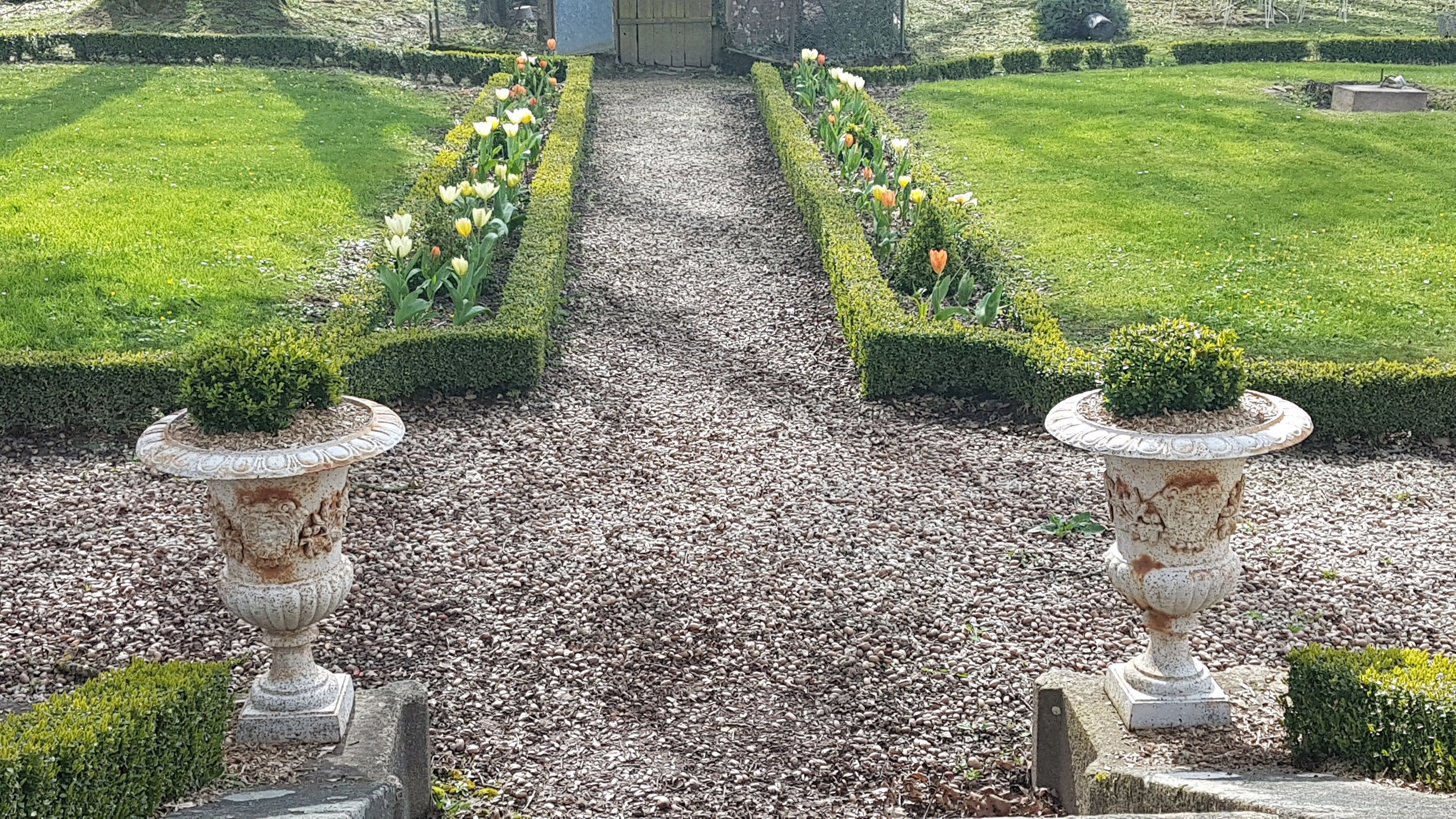 Een formele tuin met een grindpad, netjes gesnoeide hagen en twee vazen met gesnoeide planten.
