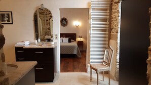 Un baño con lavabo, espejo y perchero. Se ve una cama con cabecera y mesita de noche con lámpara. Una silla es visible en la esquina.
