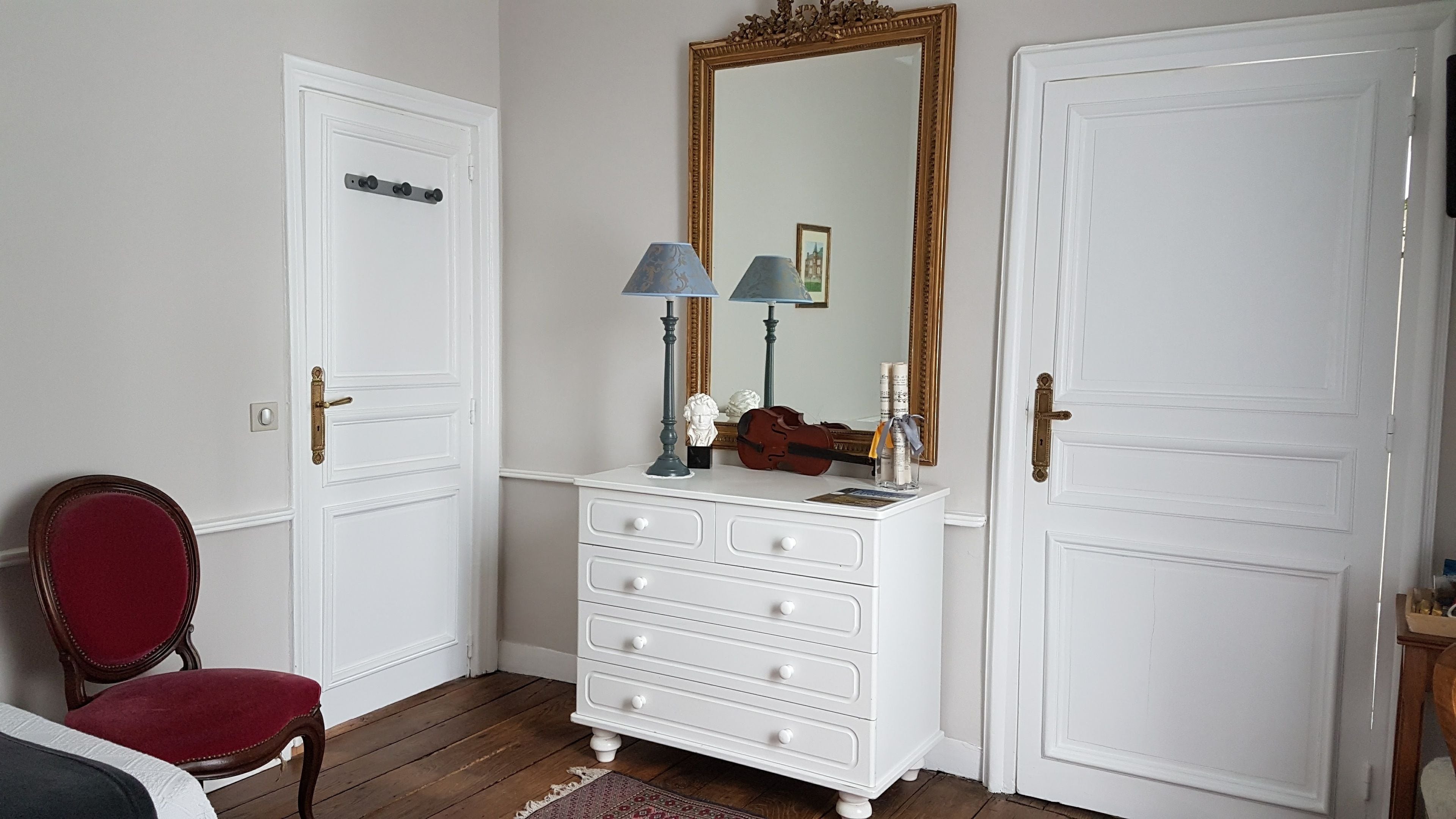 A room with a white dresser, a mirror with a gold frame, a red chair, and a violin.