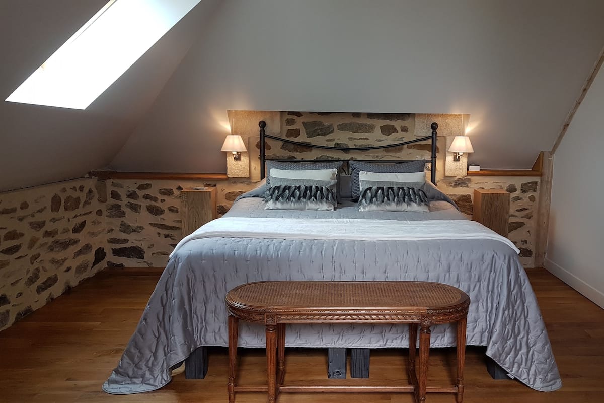 Un dormitorio con techo inclinado, paredes de piedra, una cama con ropa de cama gris, dos lámparas de noche y un banco de madera.