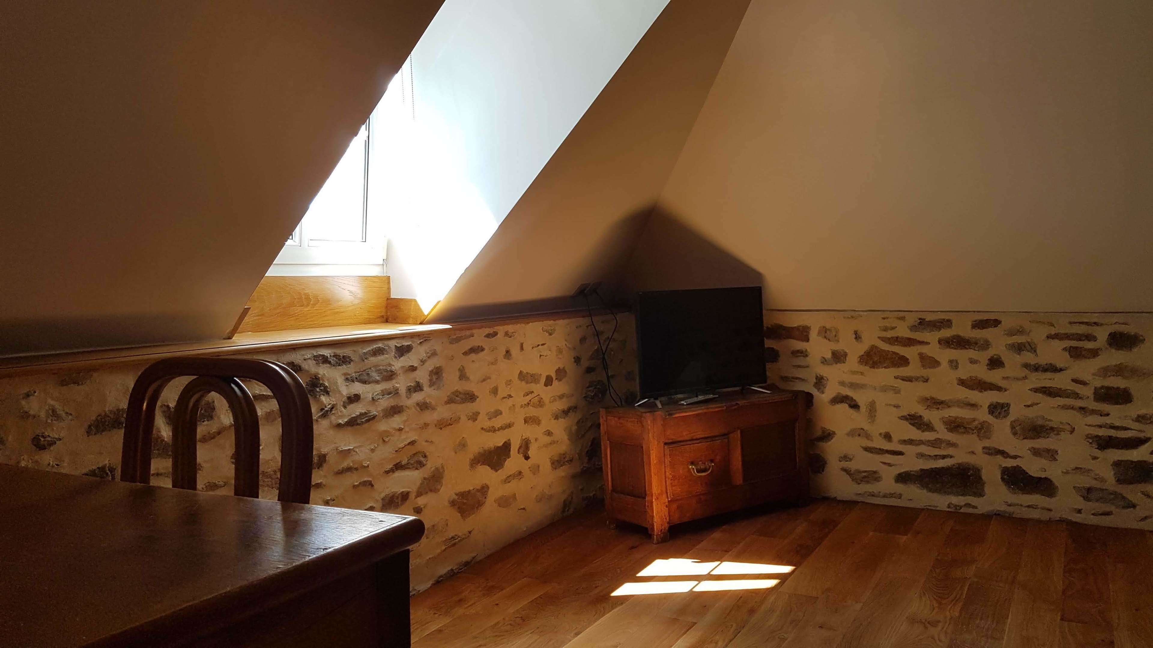 Ein kleiner, schräger Raum mit einem Holztisch, einem Stuhl, einem Fernseher auf einem Holzständer und einer Wand mit Steinmuster.