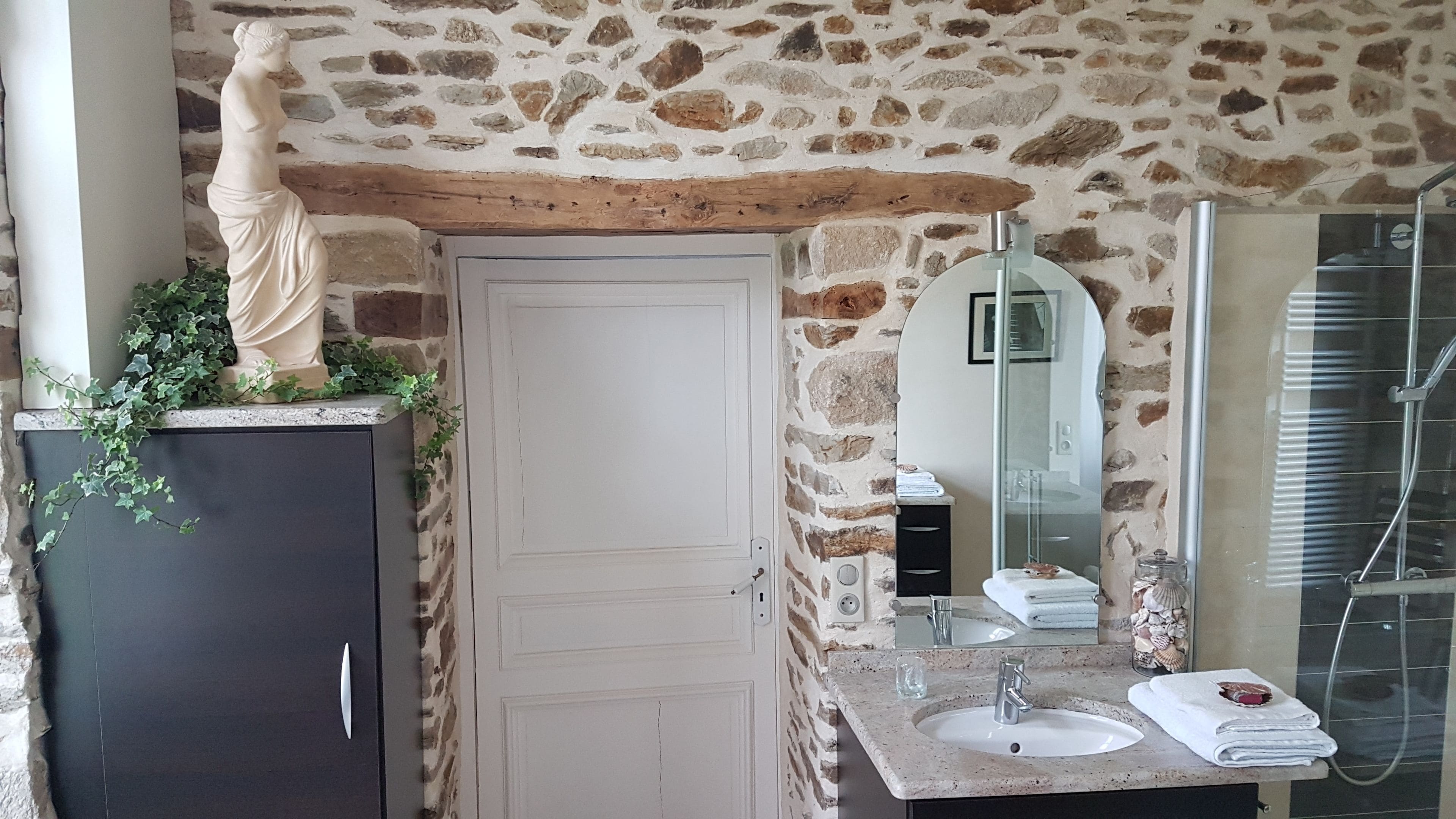 A bathroom with a stone wall, a white door, a marble sink, a mirror, and a shower.