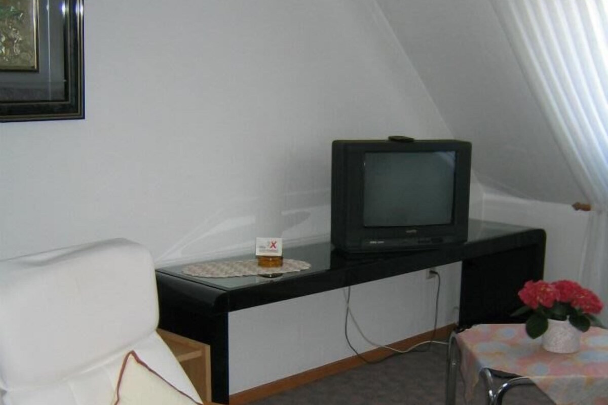 A room with a white armchair, a small table with a vase of flowers, a television on a black stand, and a framed picture on the wall.