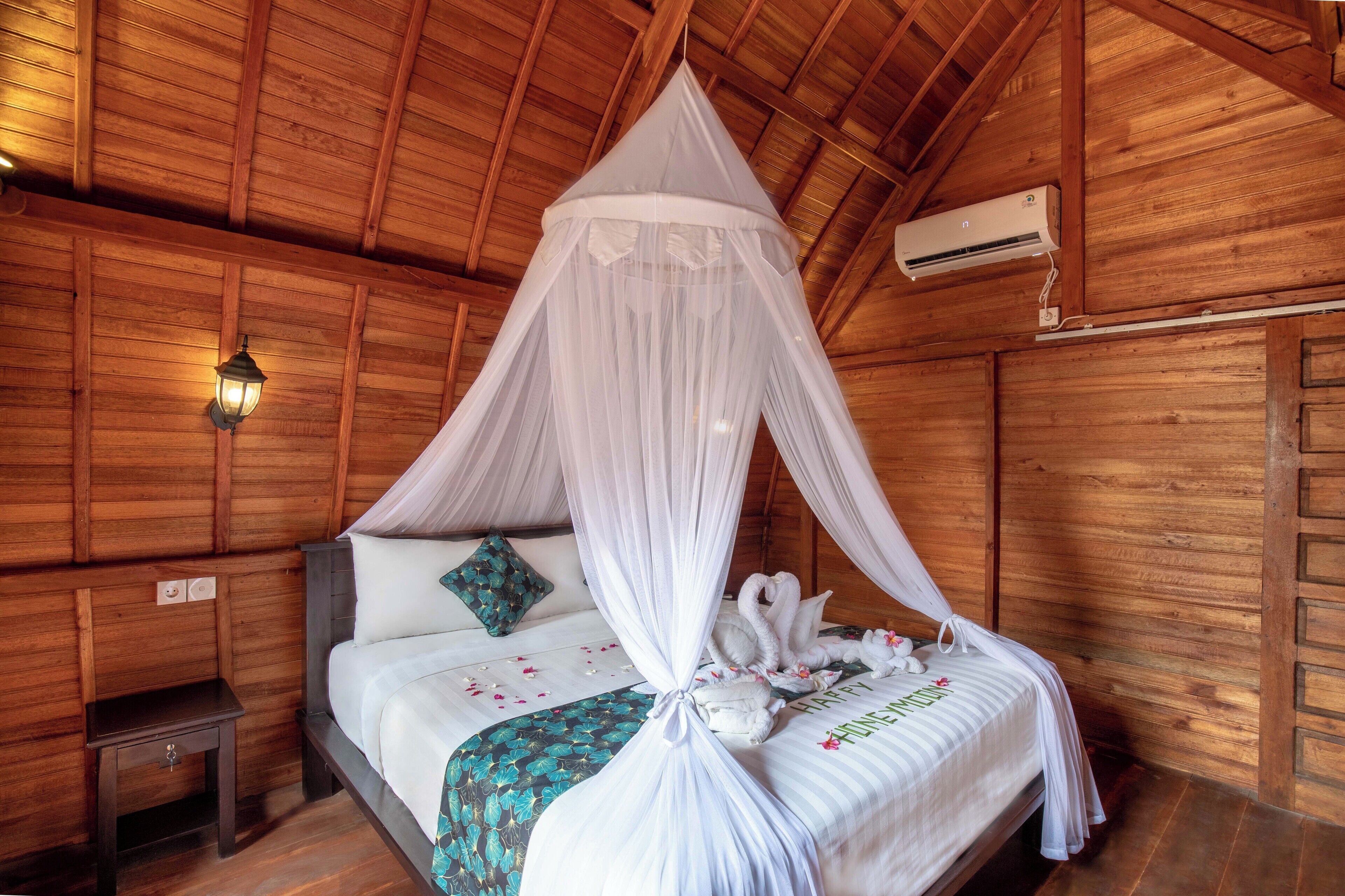 Une pièce en bois avec un lit sous un dais, une table de chevet, une lampe fixée au mur et un appareil de climatisation.