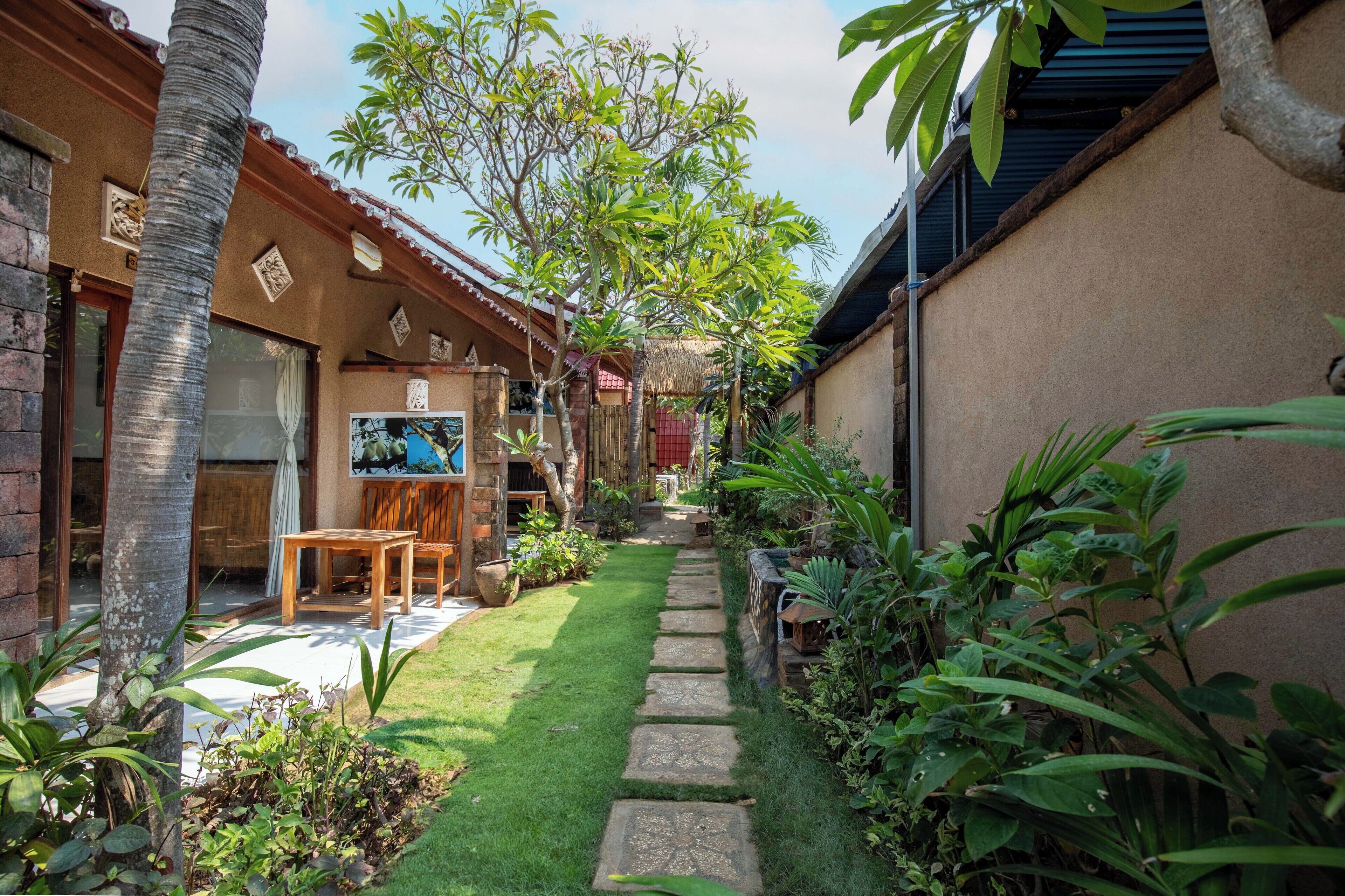A narrow pathway leading to a wooden deck with a table and chairs, surrounded by lush greenery and a thatched-roof structure.