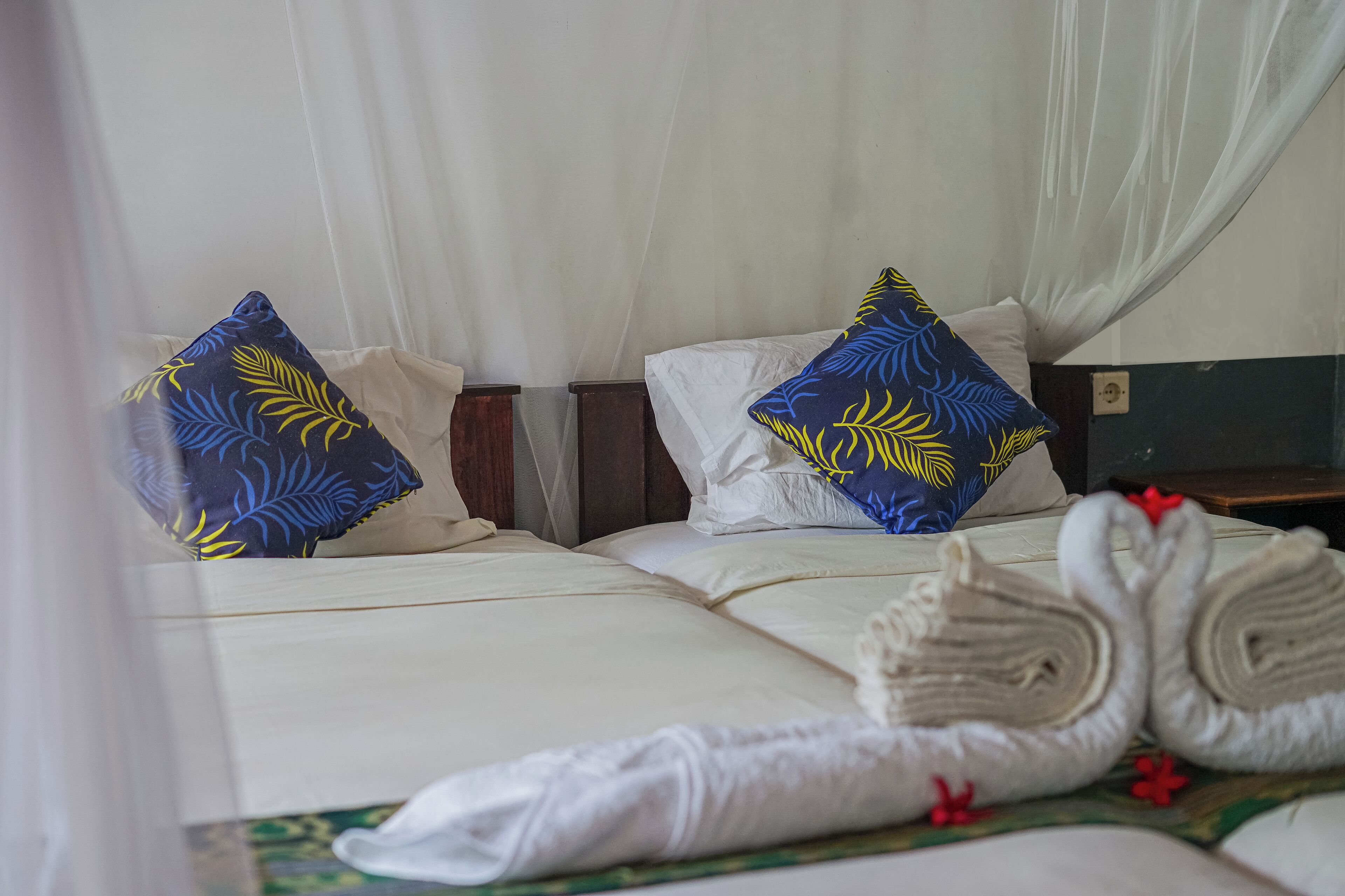 A neatly made bed with white linens and blue and yellow patterned pillows, a wooden nightstand, and a rolled white towel on the bed.