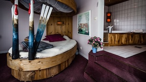 Une chambre confortable avec un lit rond en bois, une tête de lit en bois, un tableau, un vase de fleurs et une salle de bain dotée d’un meuble-lavabo en bois.