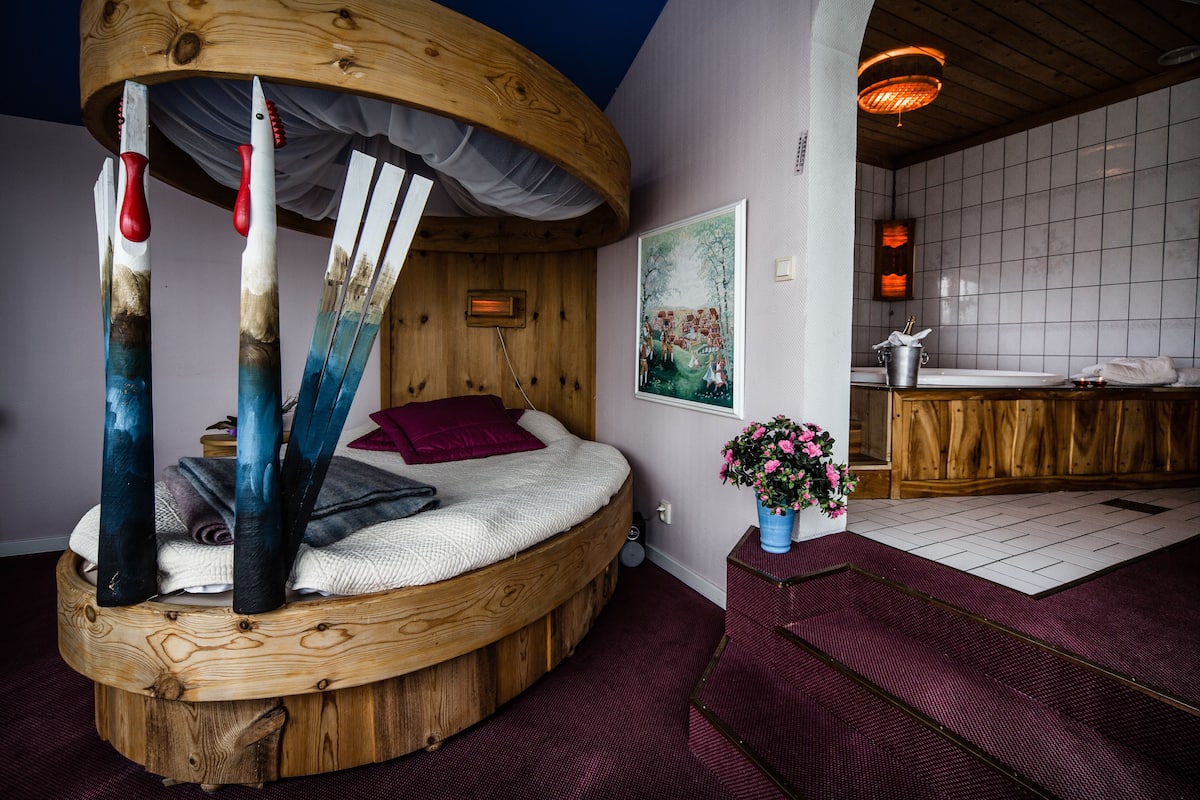 Um quarto aconchegante com uma cama redonda de madeira, cabeceira de madeira, um quadro, um vaso com flores e um banheiro com toucador de madeira.