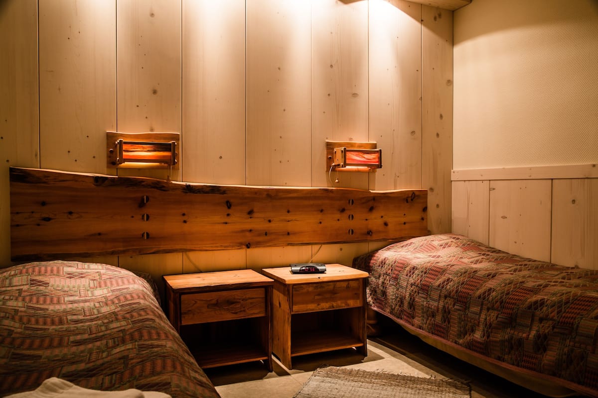 Quarto com duas camas, cabeceira de madeira, mesas de cabeceira e luminárias de parede para leitura.