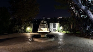 Una fuente exterior iluminada en la noche, con base circular, rodeada de un área pavimentada y luces cercanas.