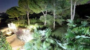 Un jardín nocturno con una fuente, árboles y senderos bien iluminados.