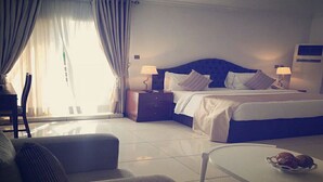 Habitación de hotel con una cama grande, dos mesitas de noche con lámparas, un sofá, una mesa de centro y un ventanal con cortinas.