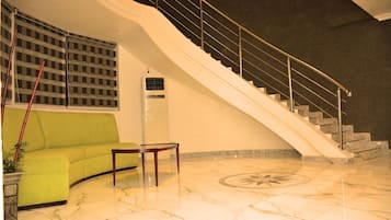 Um lobby com um sofá verde curvo, piso de mármore, escada com corrimão de metal e um aparelho de ar condicionado grande.