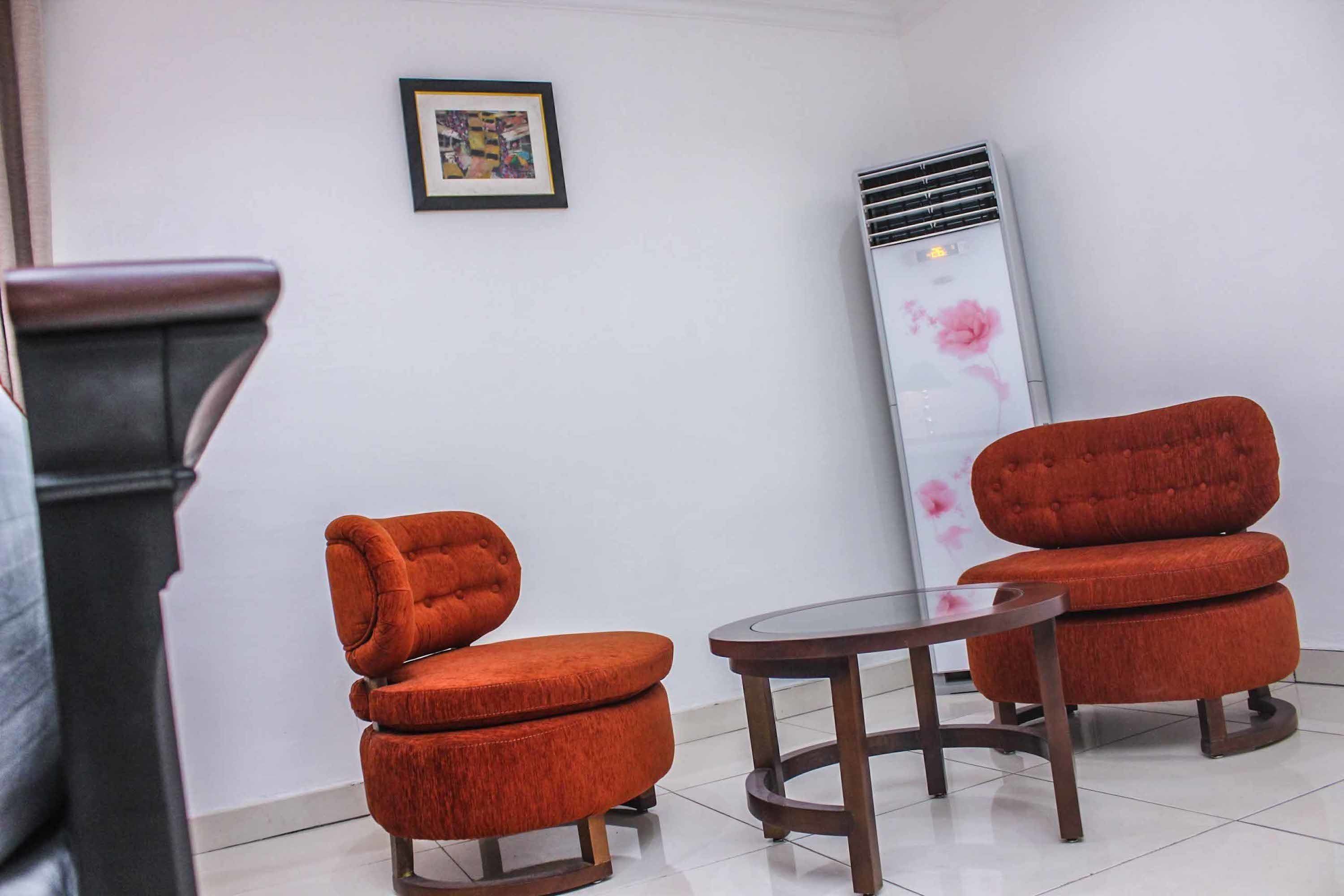 A room with two orange upholstered chairs, a small round table, a tall air conditioning unit, and a framed picture on the wall.