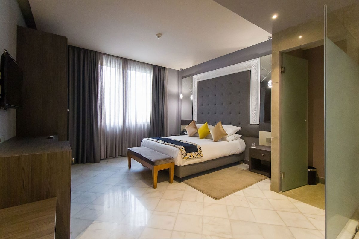 A modern hotel room with a large bed, a bedside table, a TV, and a large window with curtains.