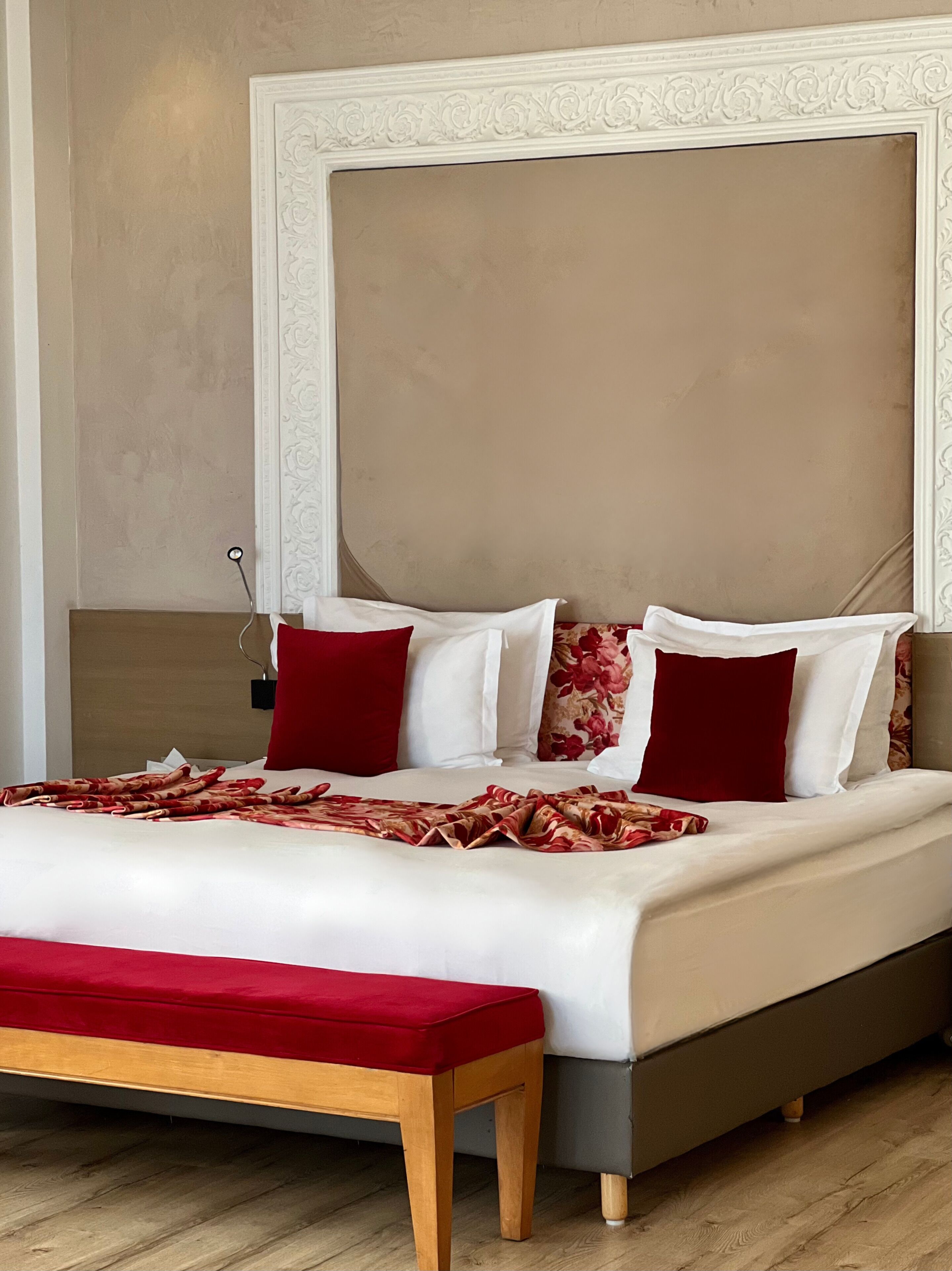 Una cama bien hecha con una colcha roja y blanca estampada, un banco de madera y un cabecero grande con un marco decorativo.