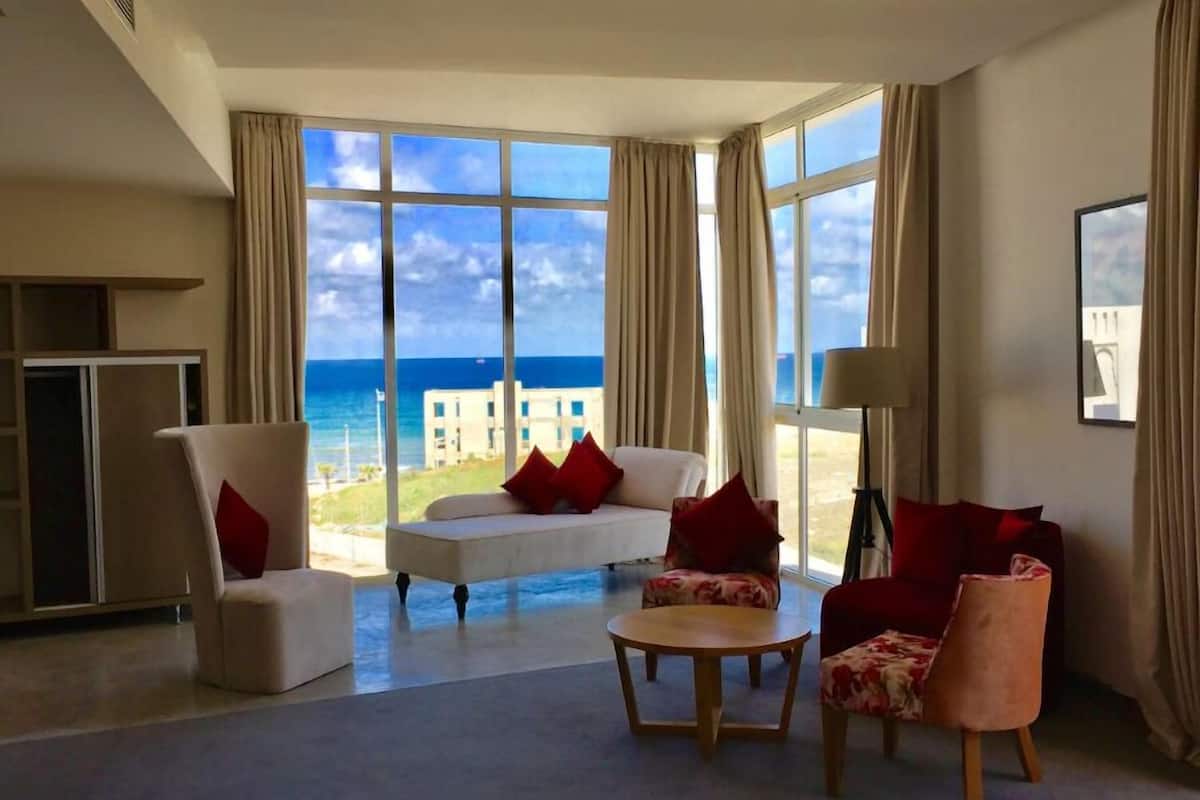 A spacious living room with a large window offering a view of the sea and a building. There are two sofas, a chair, and a small table.