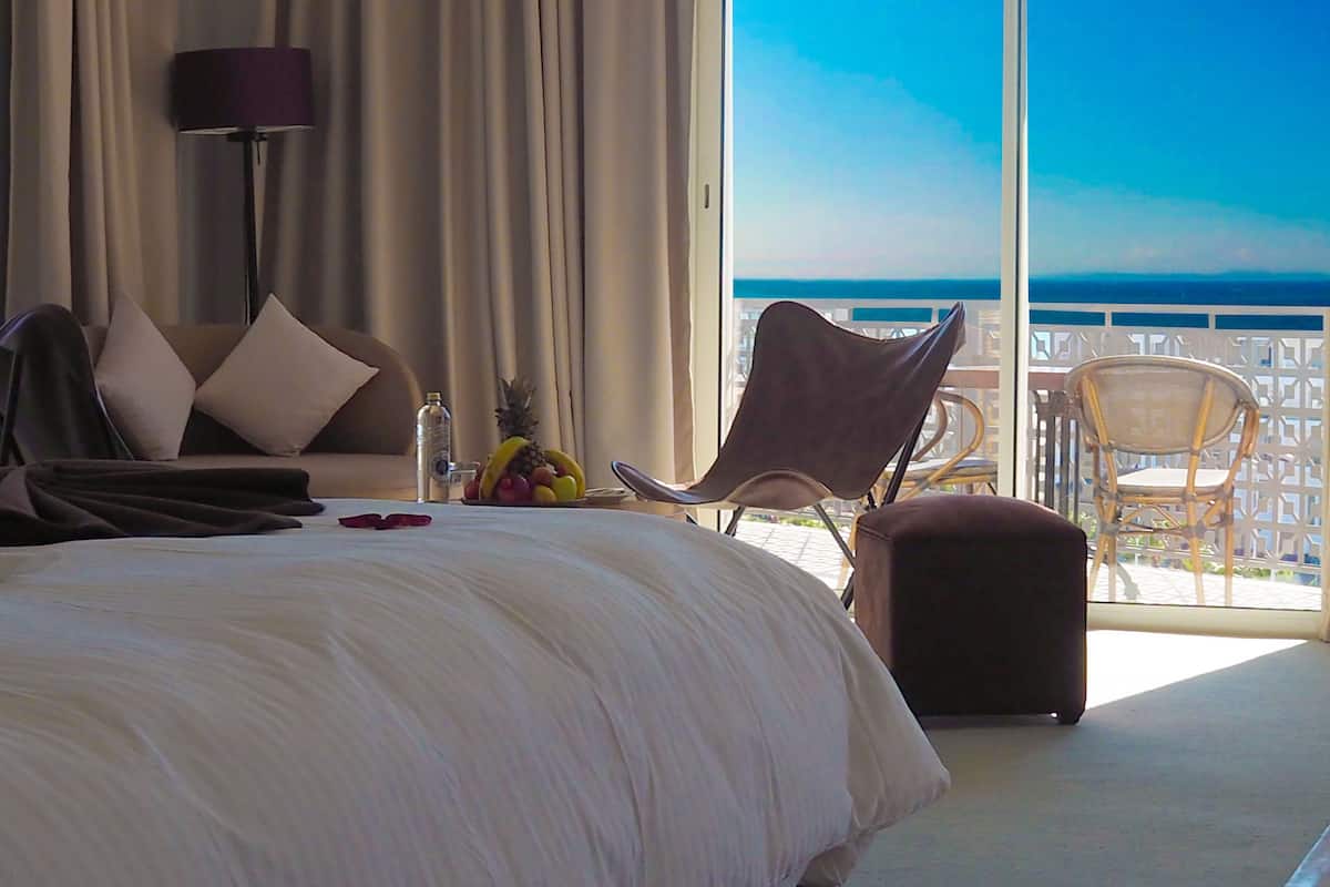 A hotel room with a bed, a sofa, a dining area with chairs, a table, and a view of the ocean.