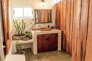 A compact bathroom with a wooden vanity, a round sink, and a mirror above it. There are potted plants on the vanity and a window above the sink. - Pura Vida Tofo Beach House (Tofo)