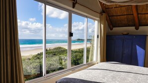 Un dormitorio con un ventanal grande con vistas a la playa, una cama con colcha estampada y techo de madera.