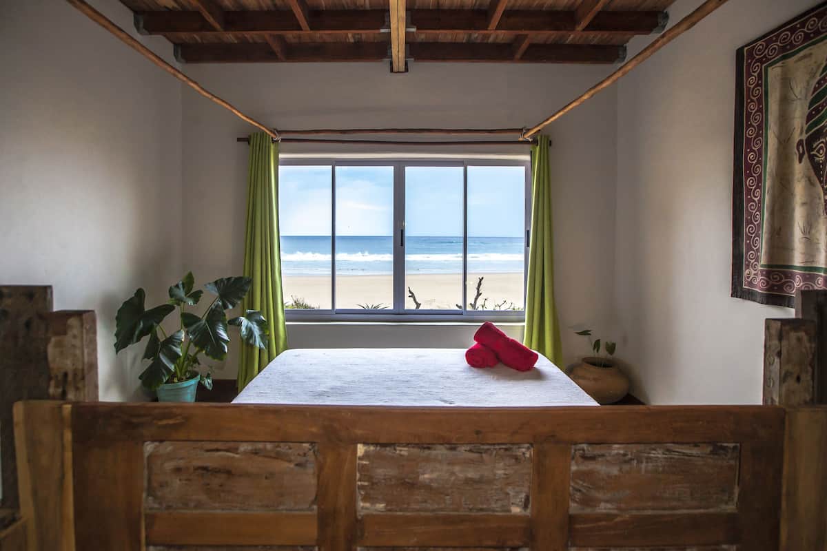 A room with a wooden bench, a table with a rolled-up red towel, a potted plant, and a view of the beach through a window with green curtains.