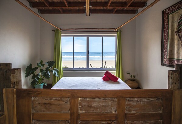 A room with a wooden bench, a table with a rolled-up red towel, a potted plant, and a view of the beach through a window with green curtains. - Pura Vida Tofo Beach House (Tofo)