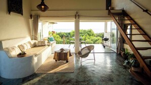 Amplia sala de estar con un sofá curvo, una mesa de centro de madera y un sillón de mimbre. Una escalera conduce a un espacio exterior con vistas a la vegetación y al mar.