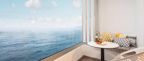 A balcony with a view of the sea, a round table with a bottle and fruits, and a sofa with cushions.