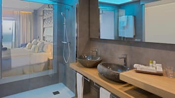 Un baño moderno con un amplio box de ducha de vidrio, un tocador de madera con un lavamanos de piedra grande y vistas a un dormitorio con cama y vista al mar.