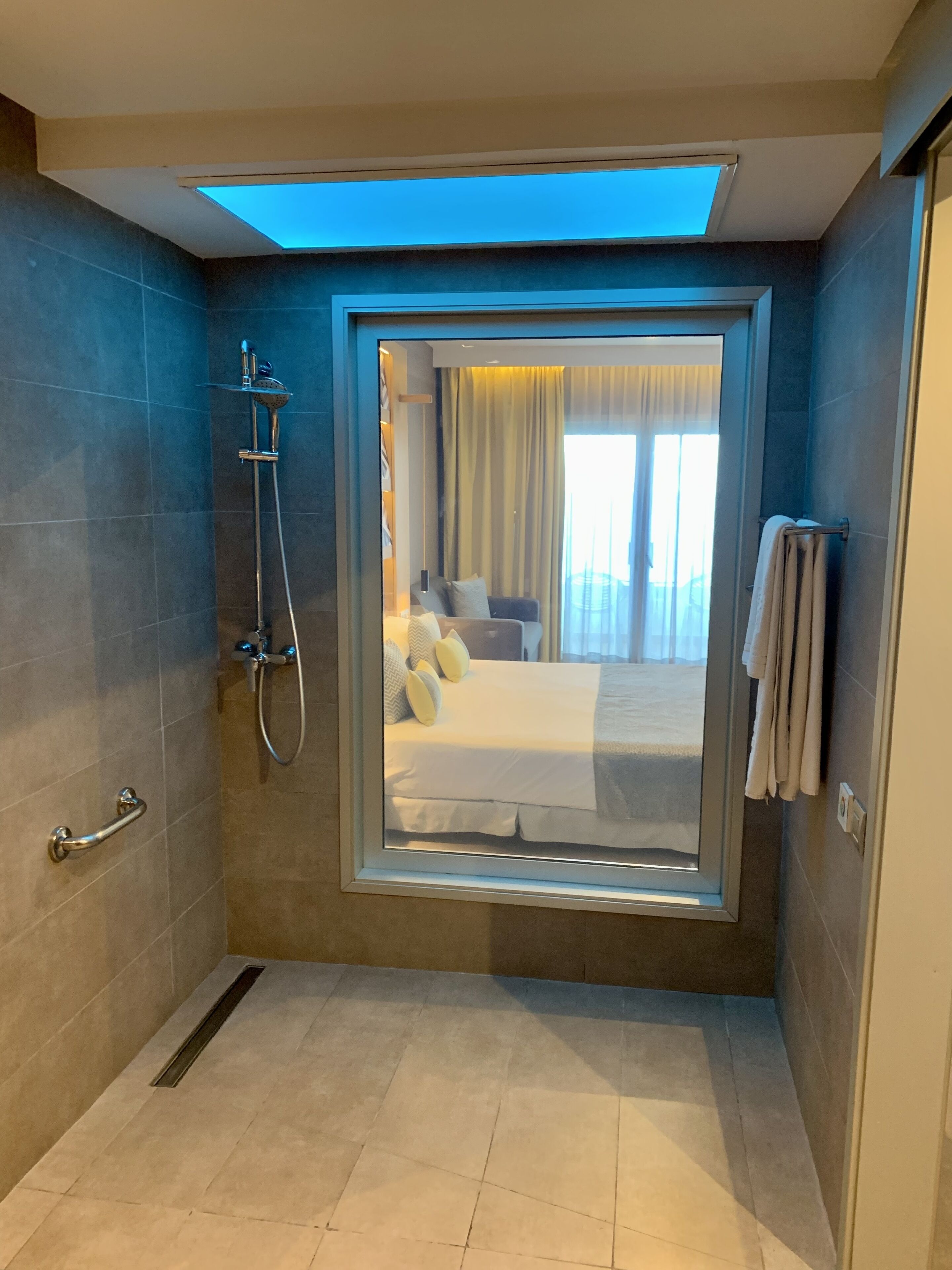 Un baño moderno con ducha de vidrio, un perchero y una habitación con cama y cortinas.
