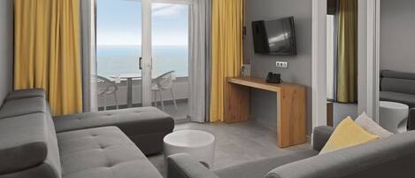 A modern living room with grey sofas, a small round table, a flat-screen TV, and a balcony with yellow curtains.