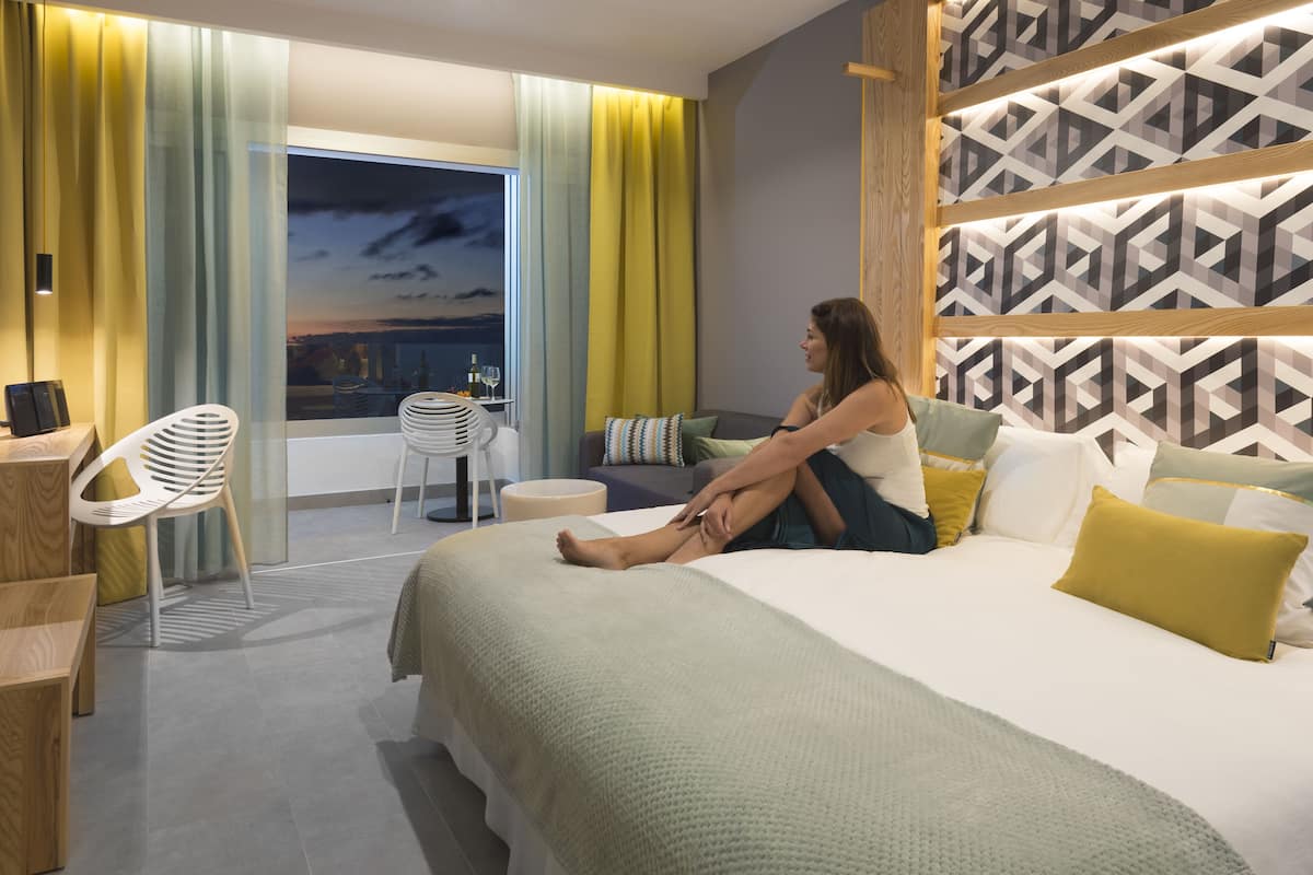 A modern hotel room with a large bed, a seating area with a chair, a small table, and a view of the city at dusk.