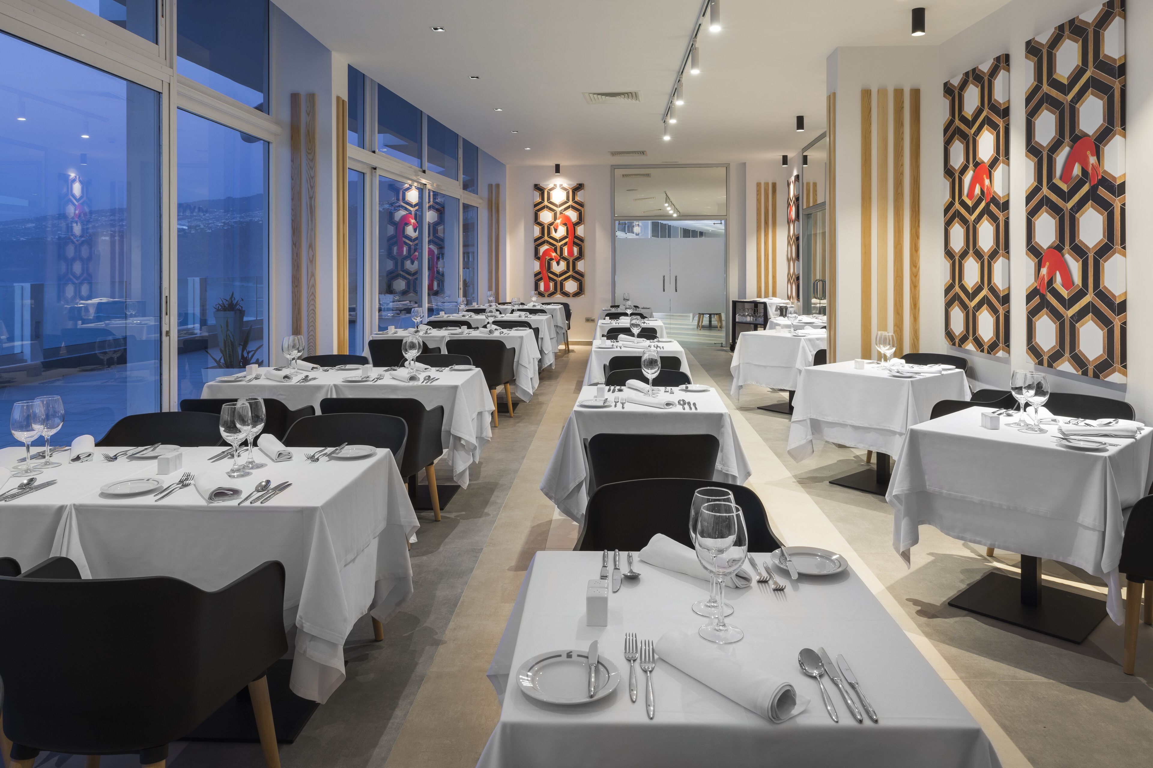 En restaurang med flera bord som är dukade för middag, stora fönster och dekorativa väggpaneler.