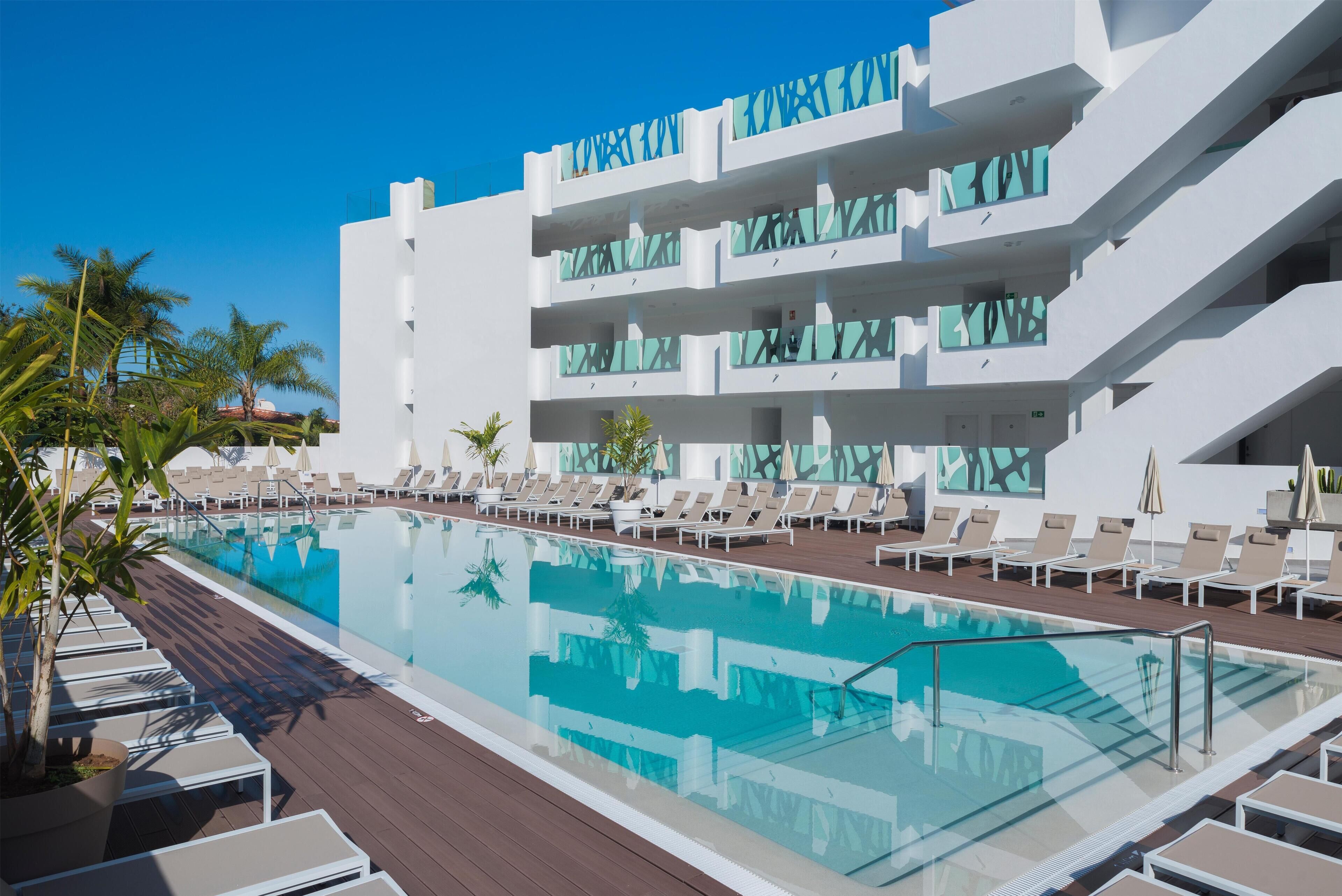 A modern hotel with a pool area, sun loungers, and a clear blue sky.