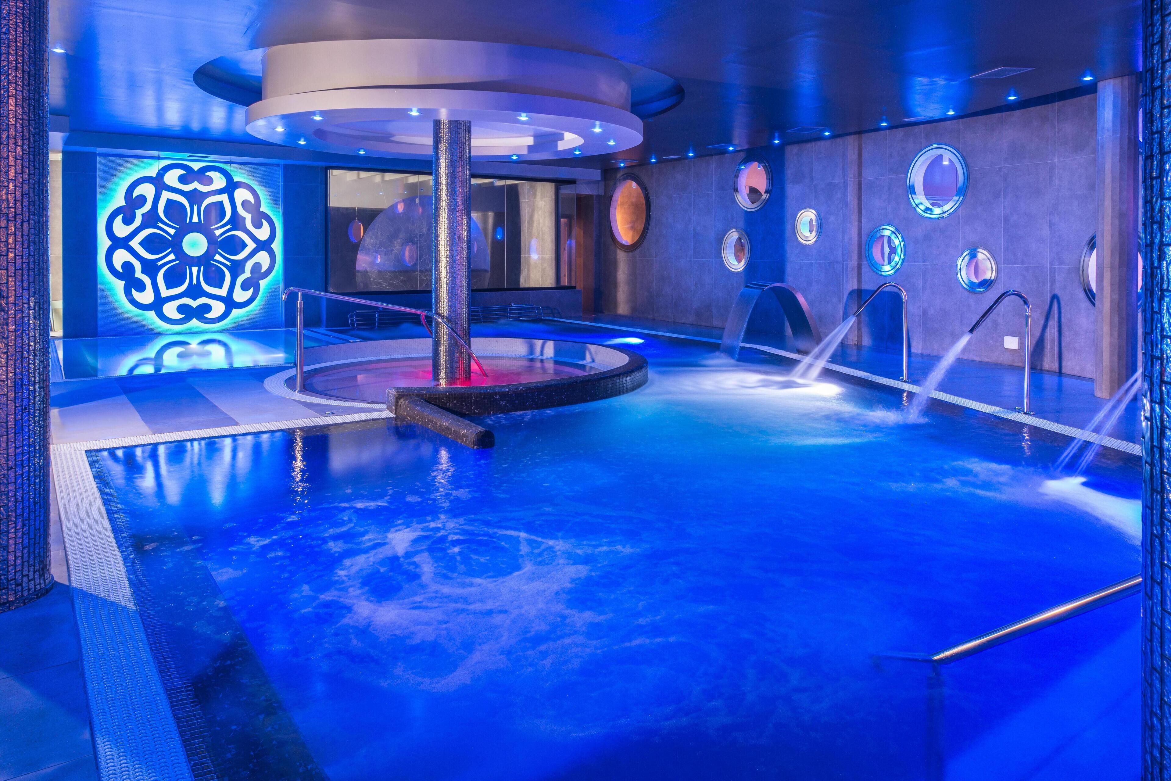 Un área de piscina interior moderna con una fuente central, varias zonas de estar y elementos decorativos en las paredes.