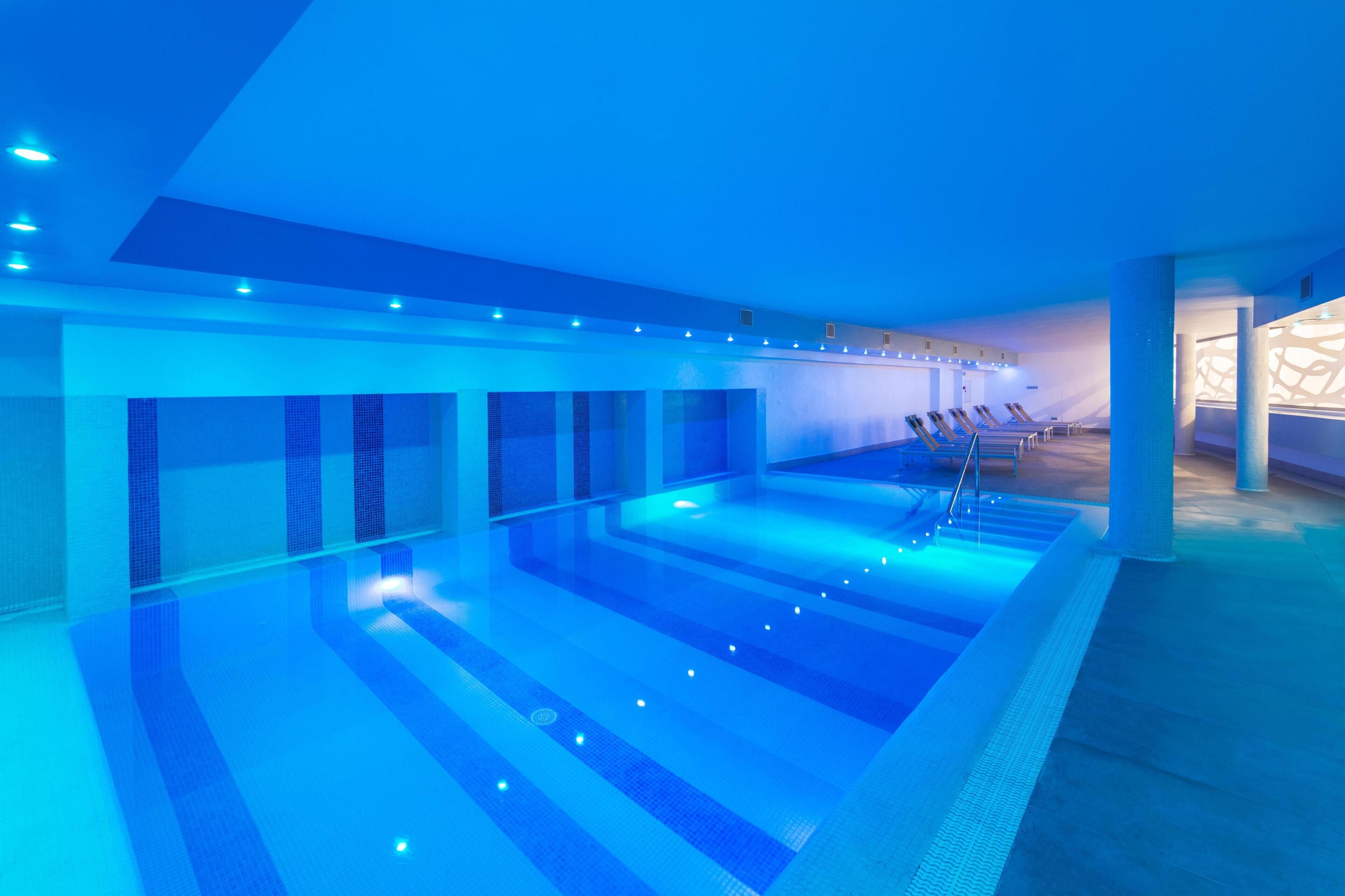 Una amplia piscina interior con iluminación azulada, múltiples carriles de natación y sillones de descanso a un lado.