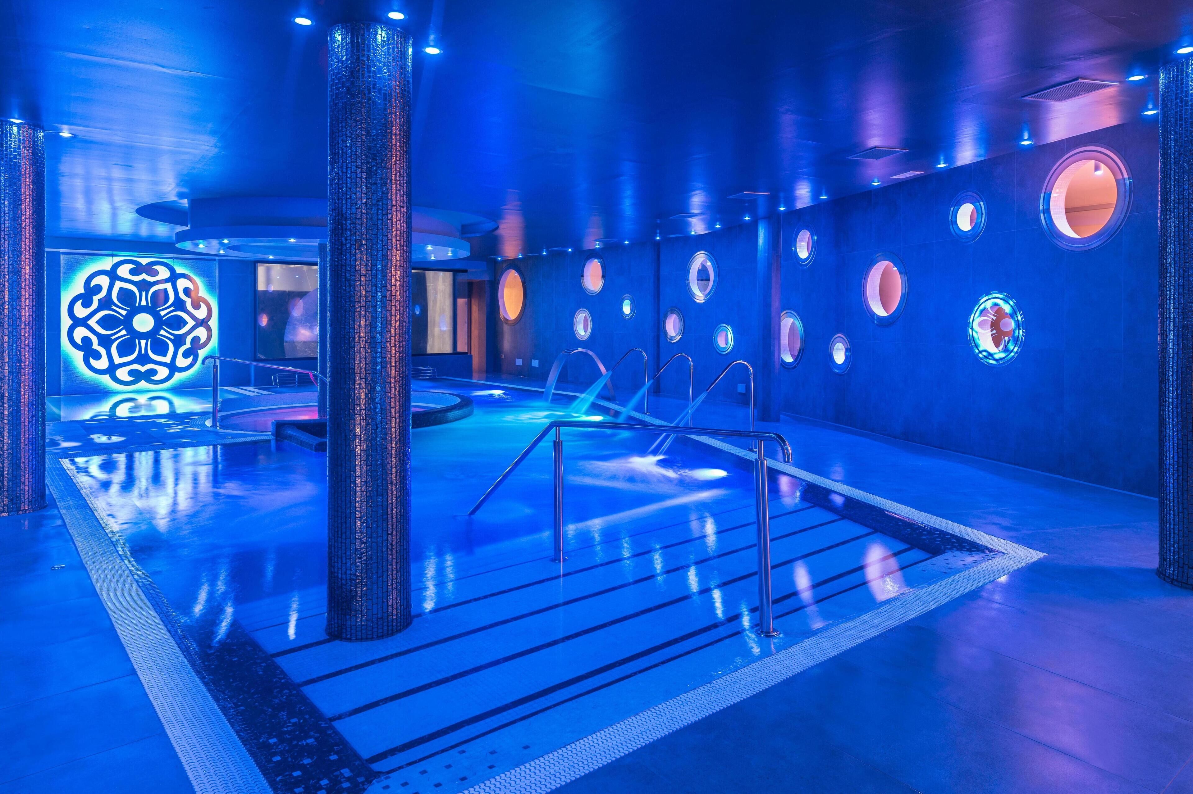 Un área de piscina interior moderna con ambiente azulado, que presenta una piscina central, varios ventanales circulares y un elemento decorativo de iluminación a la izquierda.