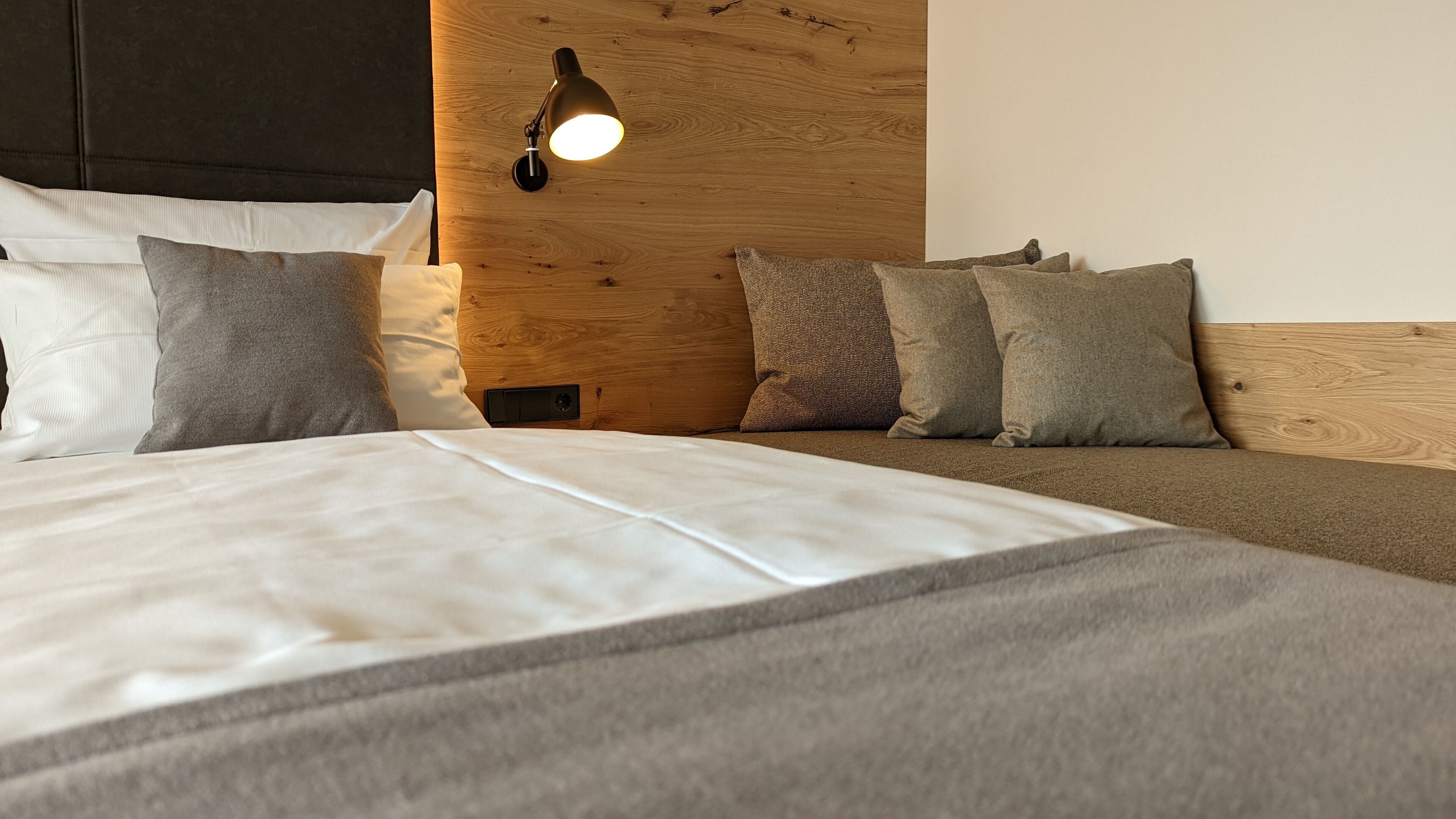 Ein ordentlich bezogenes Bett mit weißer und grauer Bettwäsche, einem hölzernen Kopfteil und einer Wandleuchte.