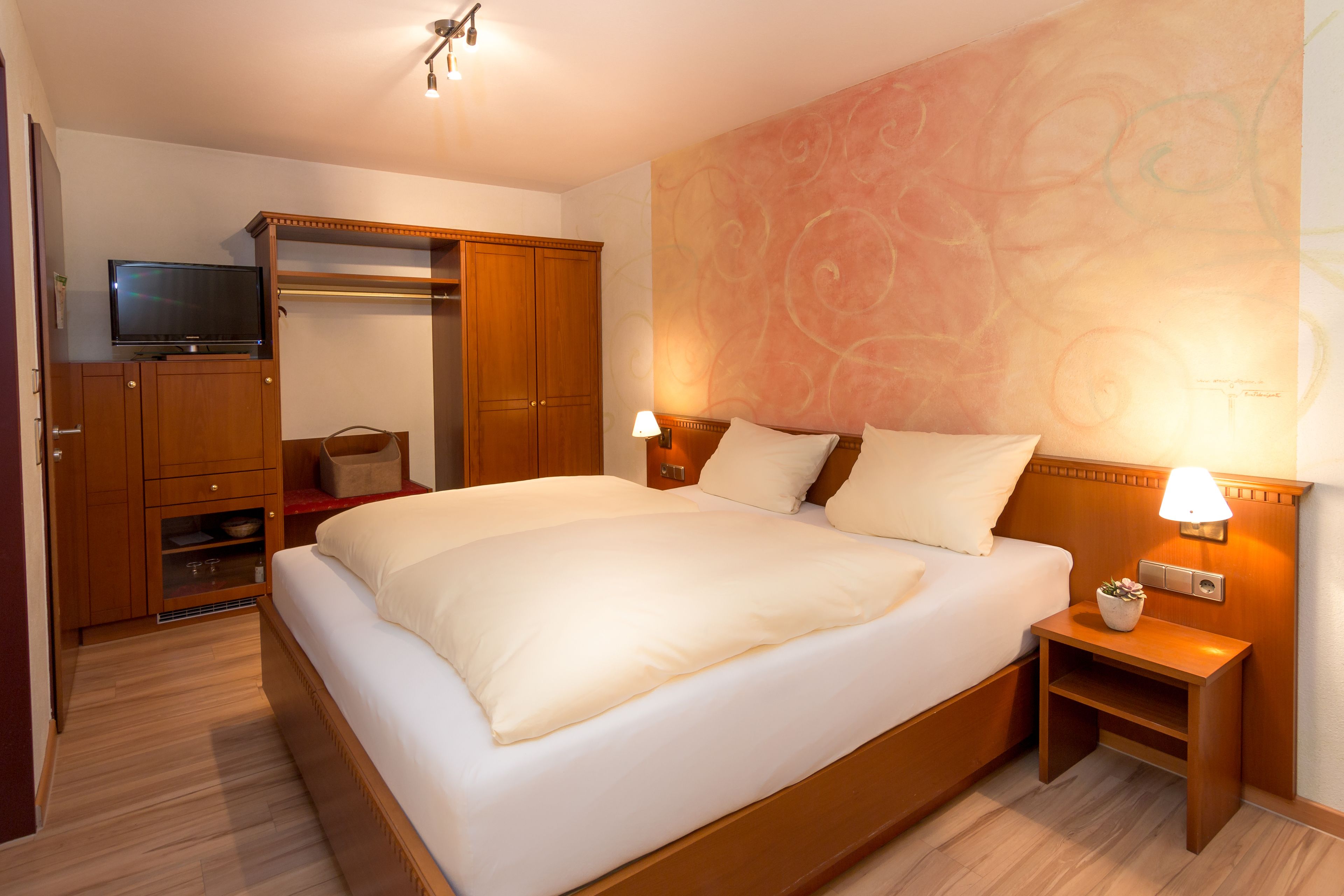 A hotel room with a large bed, wooden wardrobe, a television, and a wall with a floral pattern.
